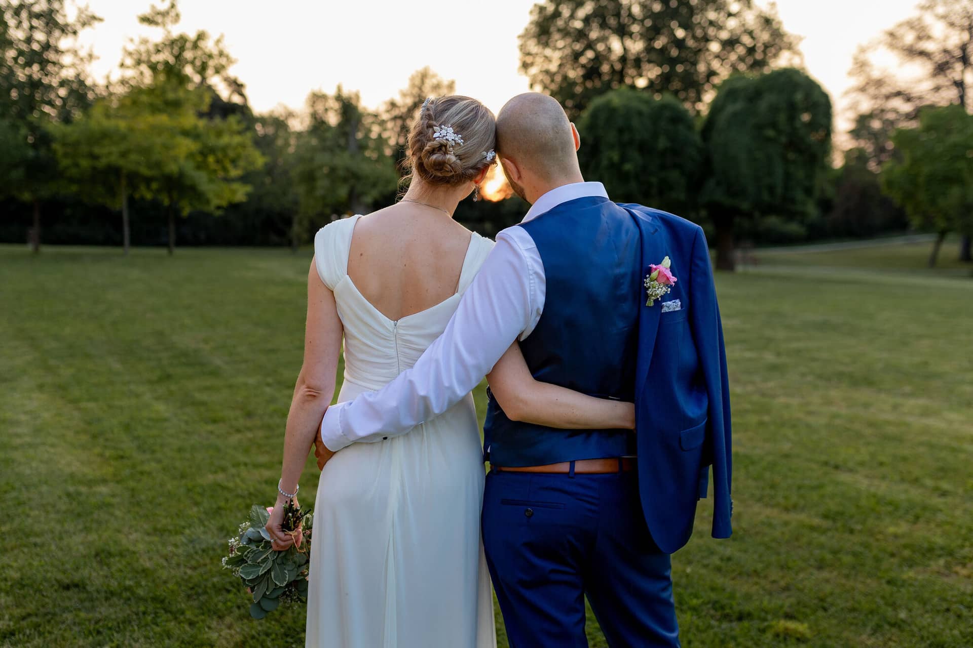 20240820 20 07 06 Teusch Schlossgarten Worms Herrnsheim 4K8A0275 Hochzeitsfotograf Matthias Butz.CZzCPH9v Brautpaar von hinten auf grüner Wiese zwischen Bäumen im Abendlicht des Schlossgartens Herrnsheim