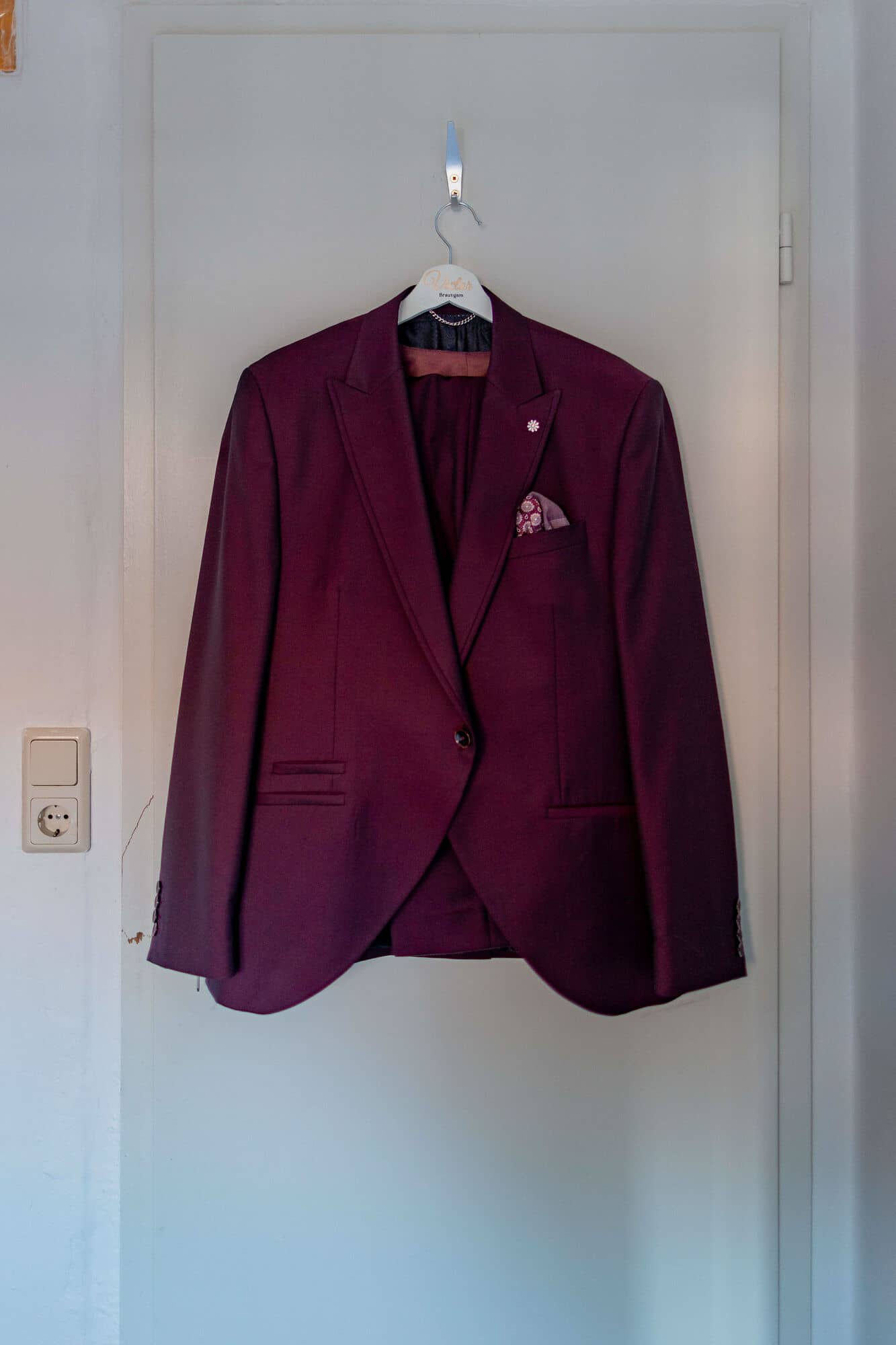 20250501 11 24 354K8A2322 Getting Ready Brautigam Bad Duerkheim.SRkjwI G Bordeauxfarbenes Hochzeitssakko hängt an weißer Tür im Hotelzimmer