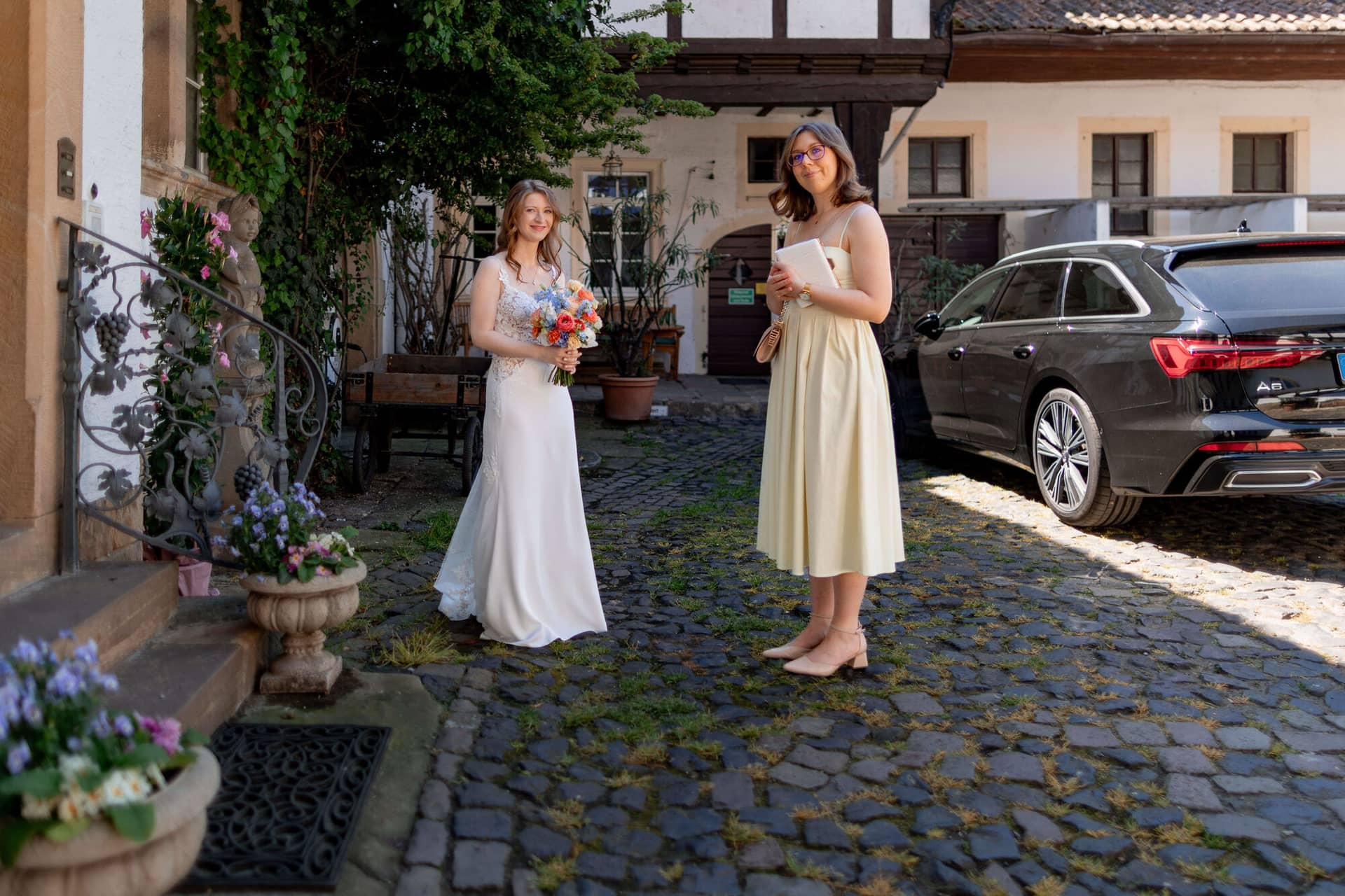 20250501 13 49 304K8A2888 First Loog Innenhof Fritz Ritter Bad Duerkheim.DM9 Vp2F Braut mit buntem Brautstrauß und Trauzeugin in beigem Kleid im Innenhof des Weinguts