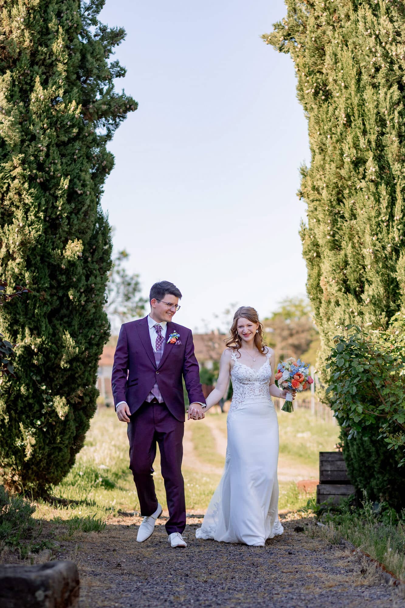 20250501 17 16 352C4A3661 Brautpaarshooting Bad Duerkheim Fritz Ritter Hochzeitsfotograf.DbueA8MM Brautpaar läuft Hand in Hand zwischen hohen Zypressen im Gutspark