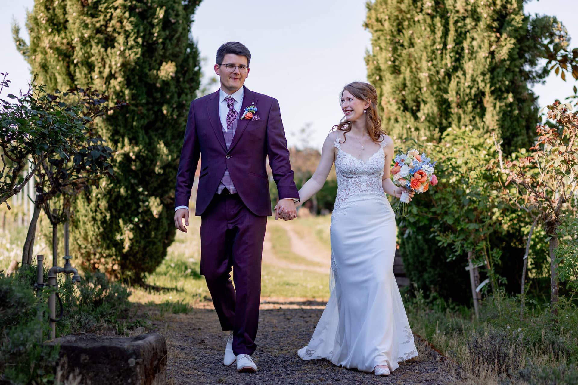 20250501 17 16 392C4A3669 Brautpaarshooting Bad Duerkheim Fritz Ritter Hochzeitsfotograf.DIRqSmFt Brautpaar spaziert entspannt zwischen Zypressen auf Kiesweg im Weingut