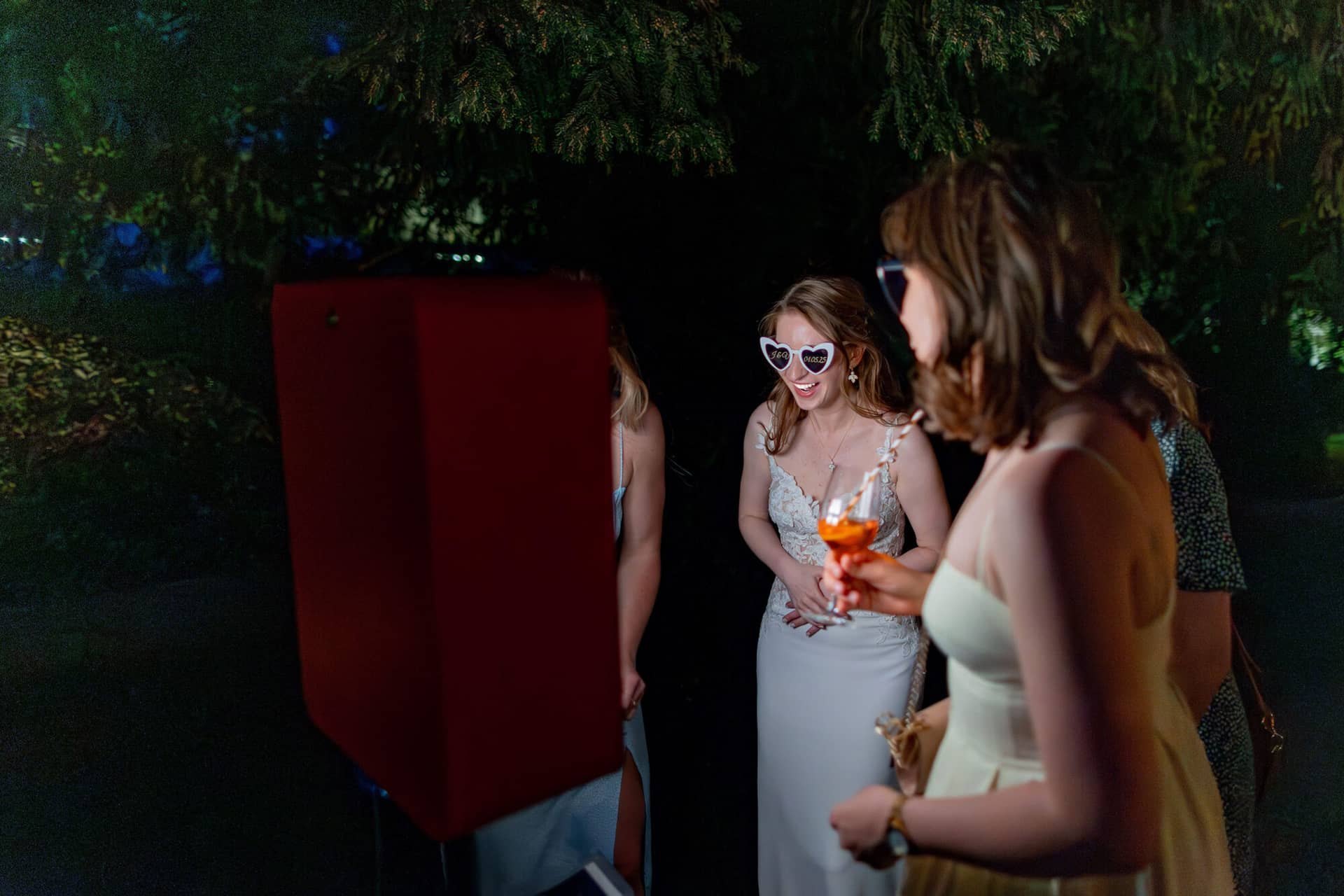 20250501 21 32 074K8A7067 Verbessert RR Hochzeitsfeier Party Bad Duerkheim Fritz Ritter Hochzeitsfotograf.BodDYi7M Braut mit herzförmiger Sonnenbrille und Gästen vor roter Fotobox bei Nacht