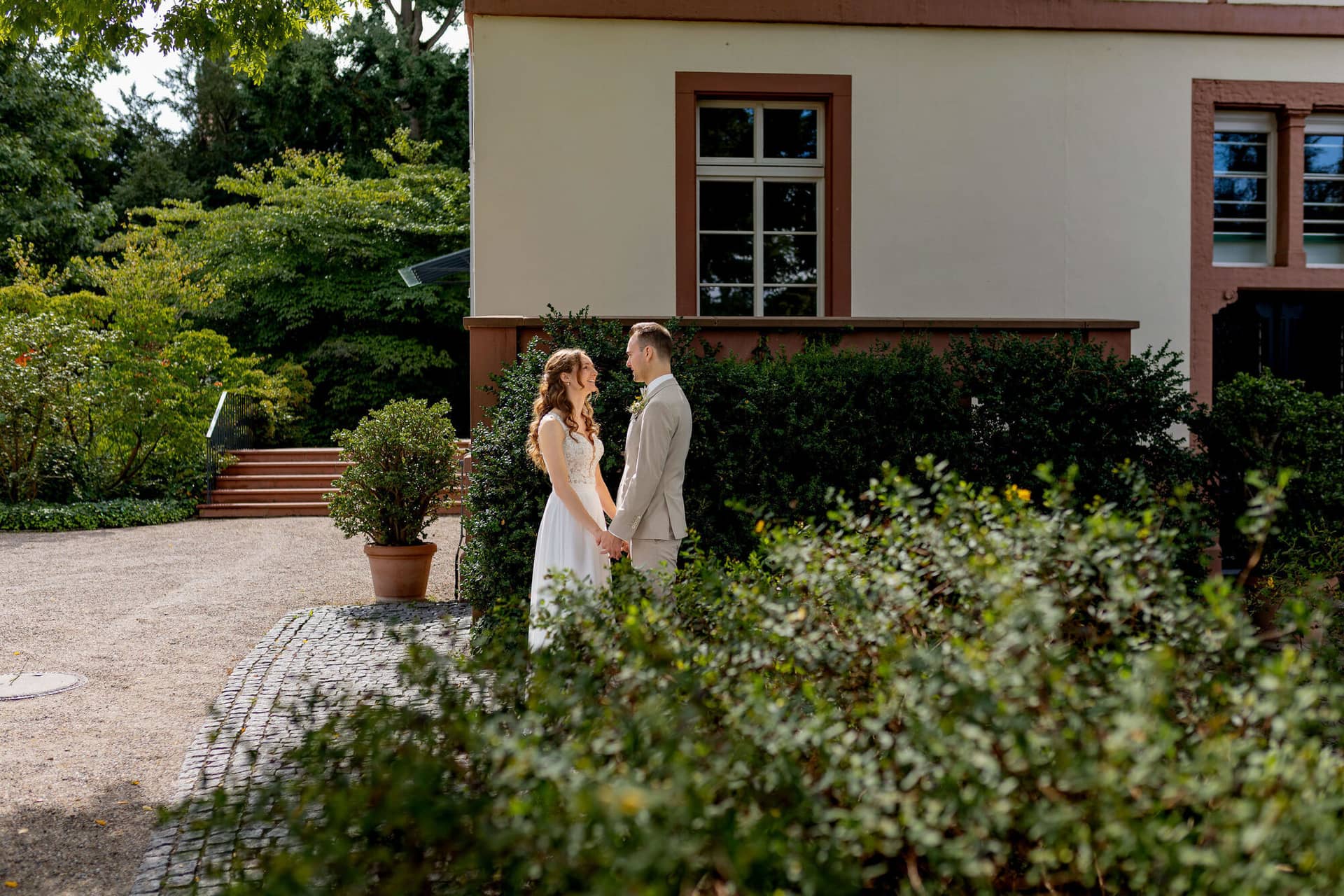 20250830 12 39 54 4K8A9879.C0ALCGpZ Brautpaar steht sich gegenüber vor historischem Gebäude mit Hecke