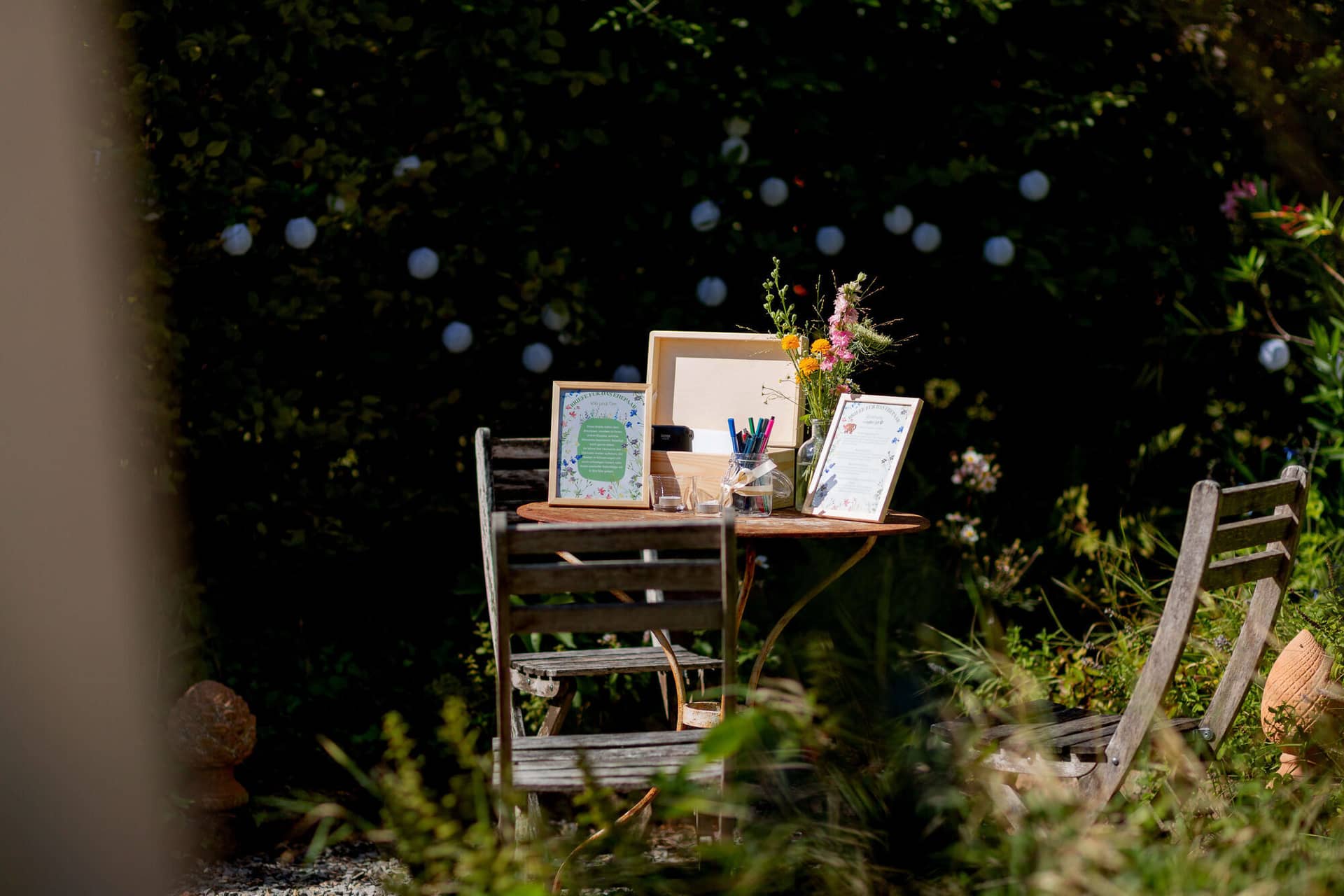 20250830 15 12 47 MK3 1115.B9I1 YrE Kleiner Holztisch mit Bilderrahmen, Stiften und Blumen im grünen Garten