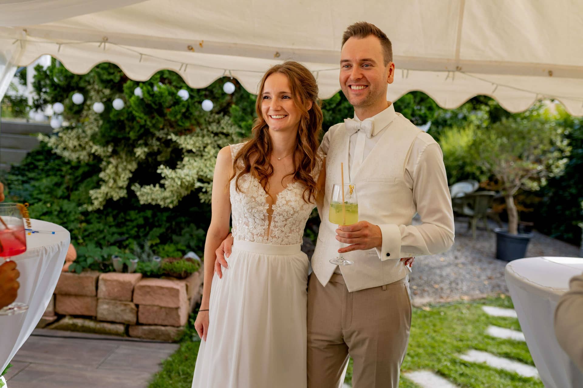 20250830 16 39 12 4K8A2150.CrSkDdbr Brautpaar lächelt in die Kamera mit Cocktails bei der Gartenparty