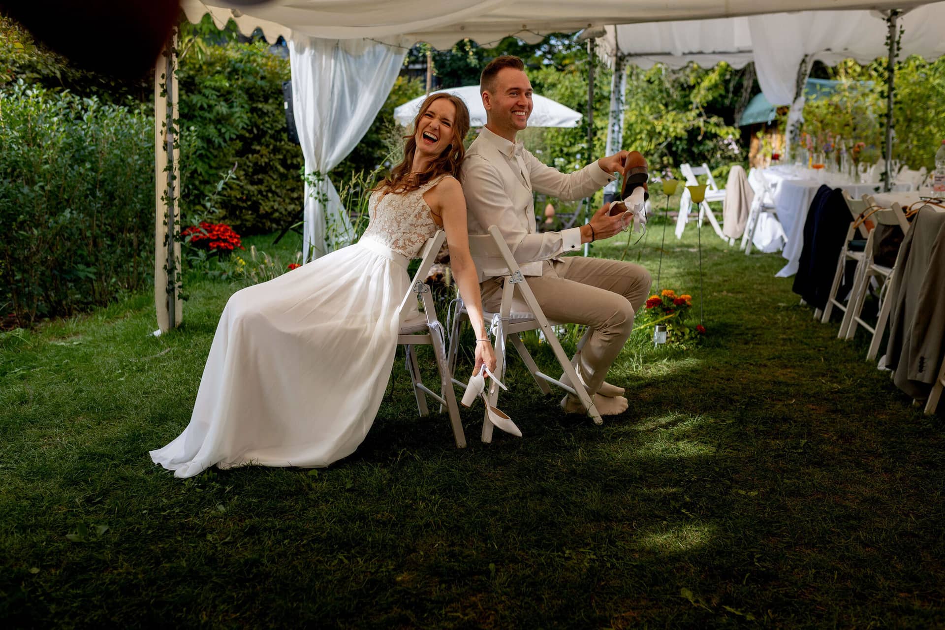 20250830 17 12 37 4K8A2695.BPSY3cqq Brautpaar sitzt lachend beim Schuhspiel auf Klappstühlen im Garten