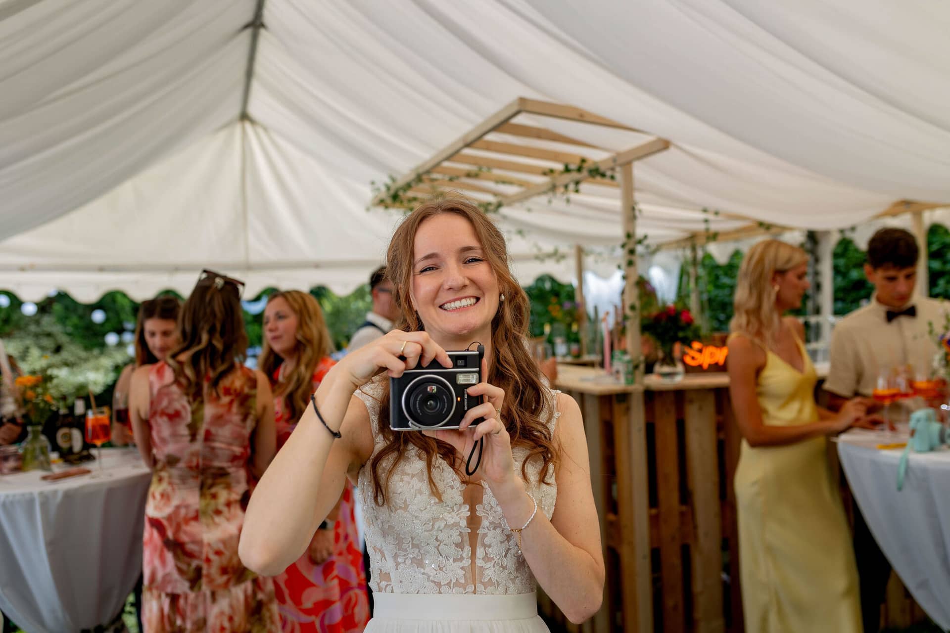 20250830 17 19 45 4K8A2746.BpfrhTUM Braut hält lächelnd eine Instax-Kamera im geschmückten Zelt