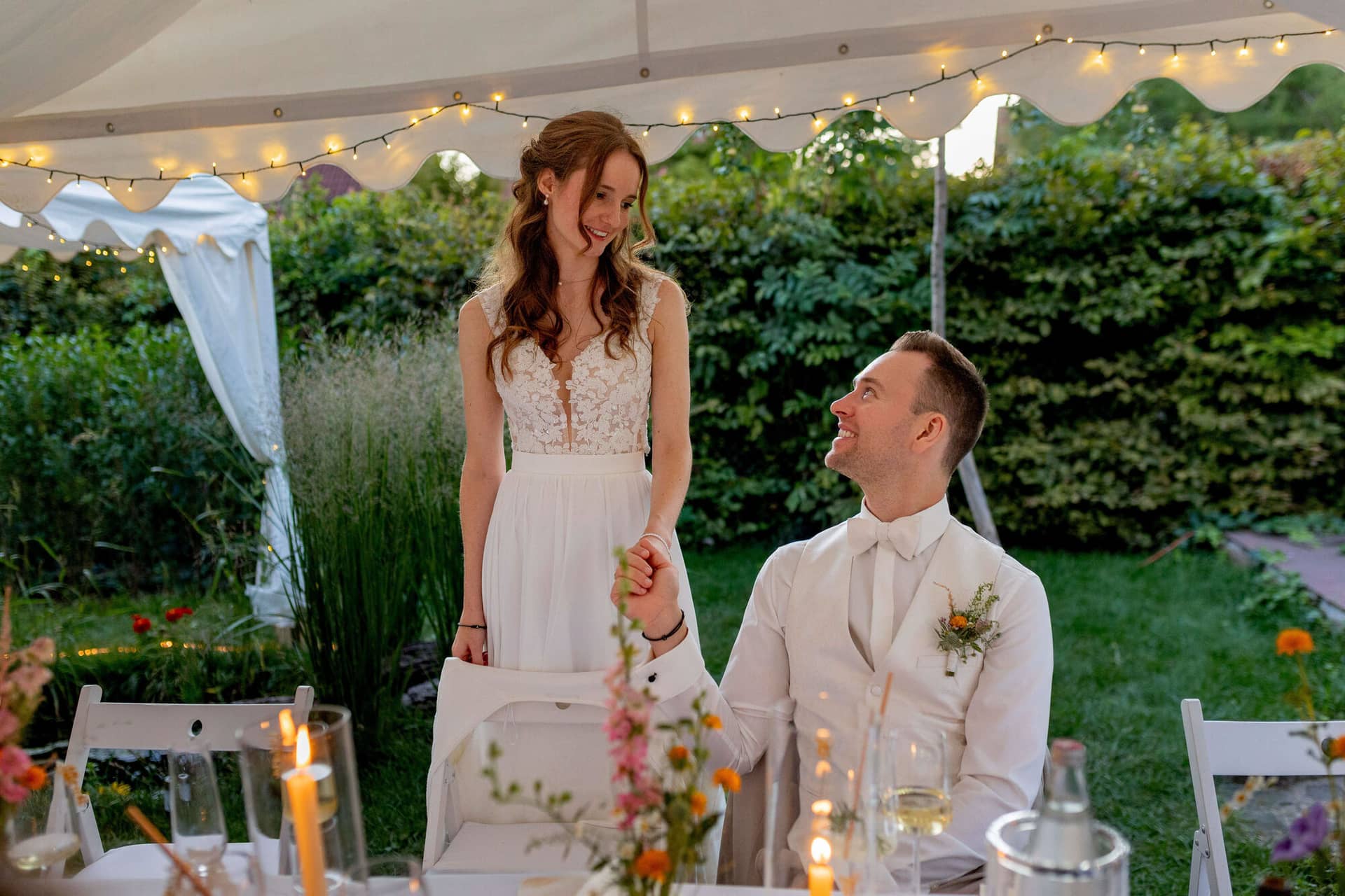 20250830 20 08 12 4K8A3417.CRQxMbnJ Braut steht lachend neben dem sitzenden Bräutigam im beleuchteten Zelt