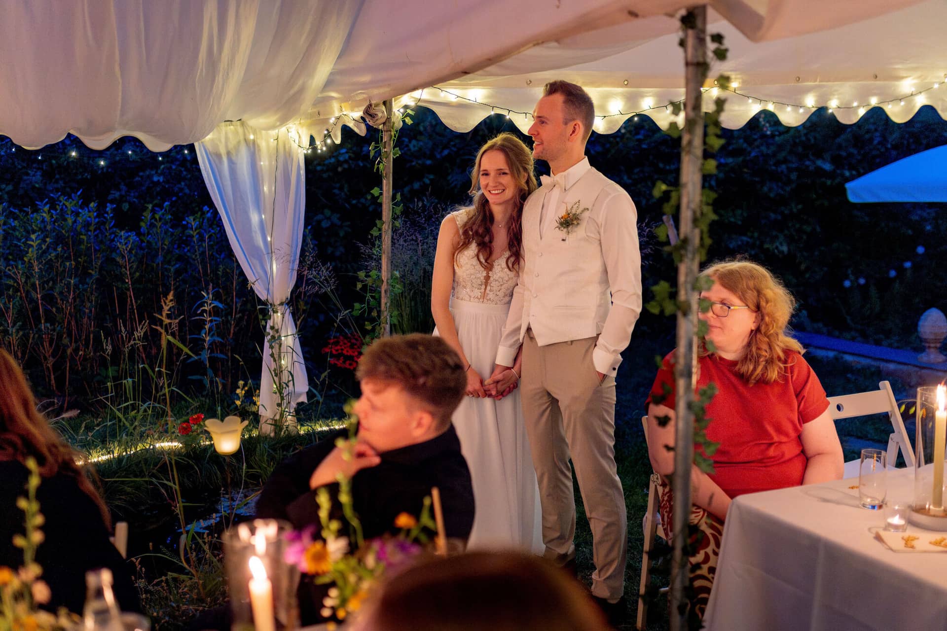 20250830 20 43 28 4K8A3527.Dp 5si1X Brautpaar steht händchenhaltend vor den Gästen im beleuchteten Zelt am Abend