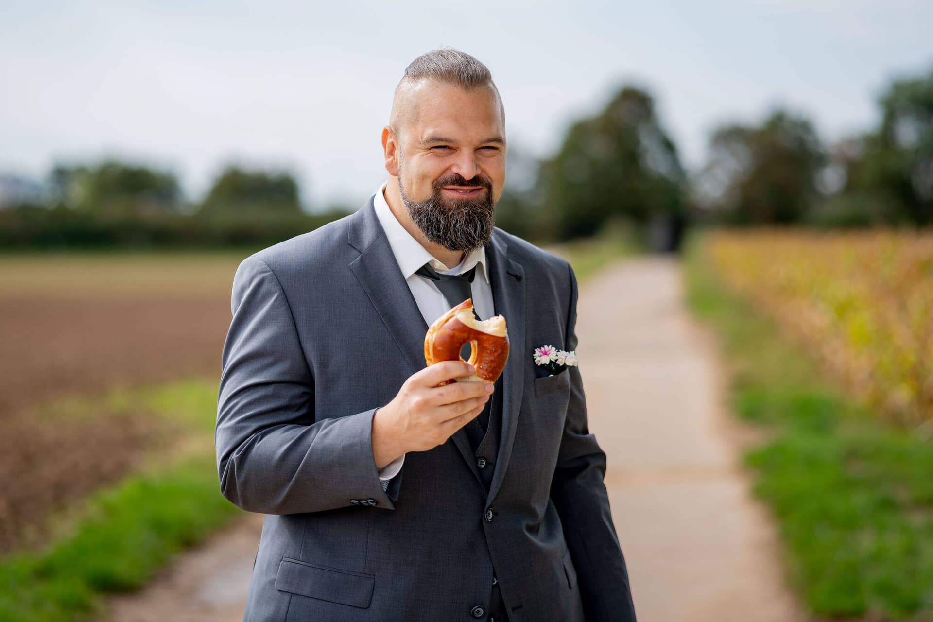 20250913 11 13 26 DSC04431.Bb9xsZf0 Bräutigam im grauen Anzug isst eine Brezel auf einem Feldweg
