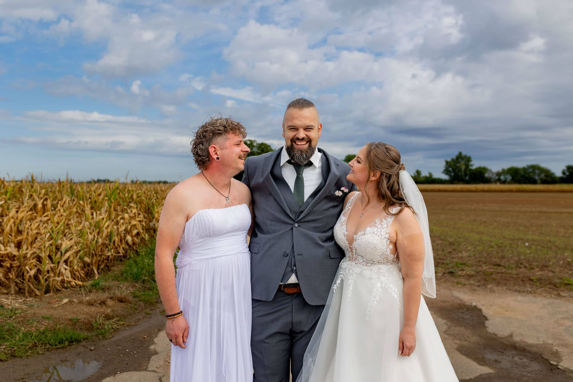 20250913 11 34 05 4K8A4393.DVkmwovq Bräutigam steht lachend zwischen Freund im weißen Kleid und Braut vor Kornfeld