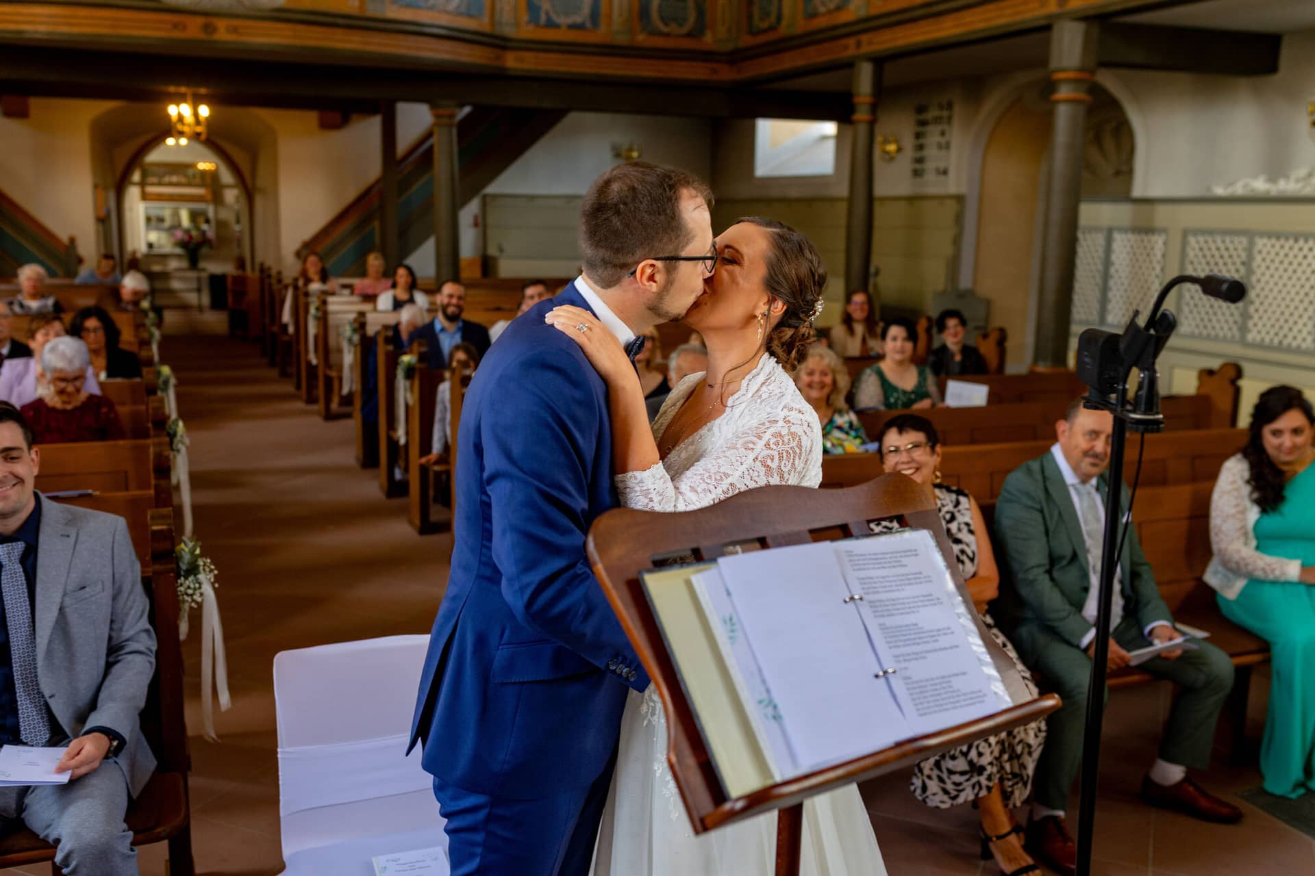 3K8A8695 Hochzeitsfotograf Mutterstadt Grenzhof Heidelberg .Ccz C X6 Braut und Bräutigam küssen sich beim ersten Kuss vor dem Hochzeitsdokumenten-Stand in der voll besetzten Kirche Mutterstadt