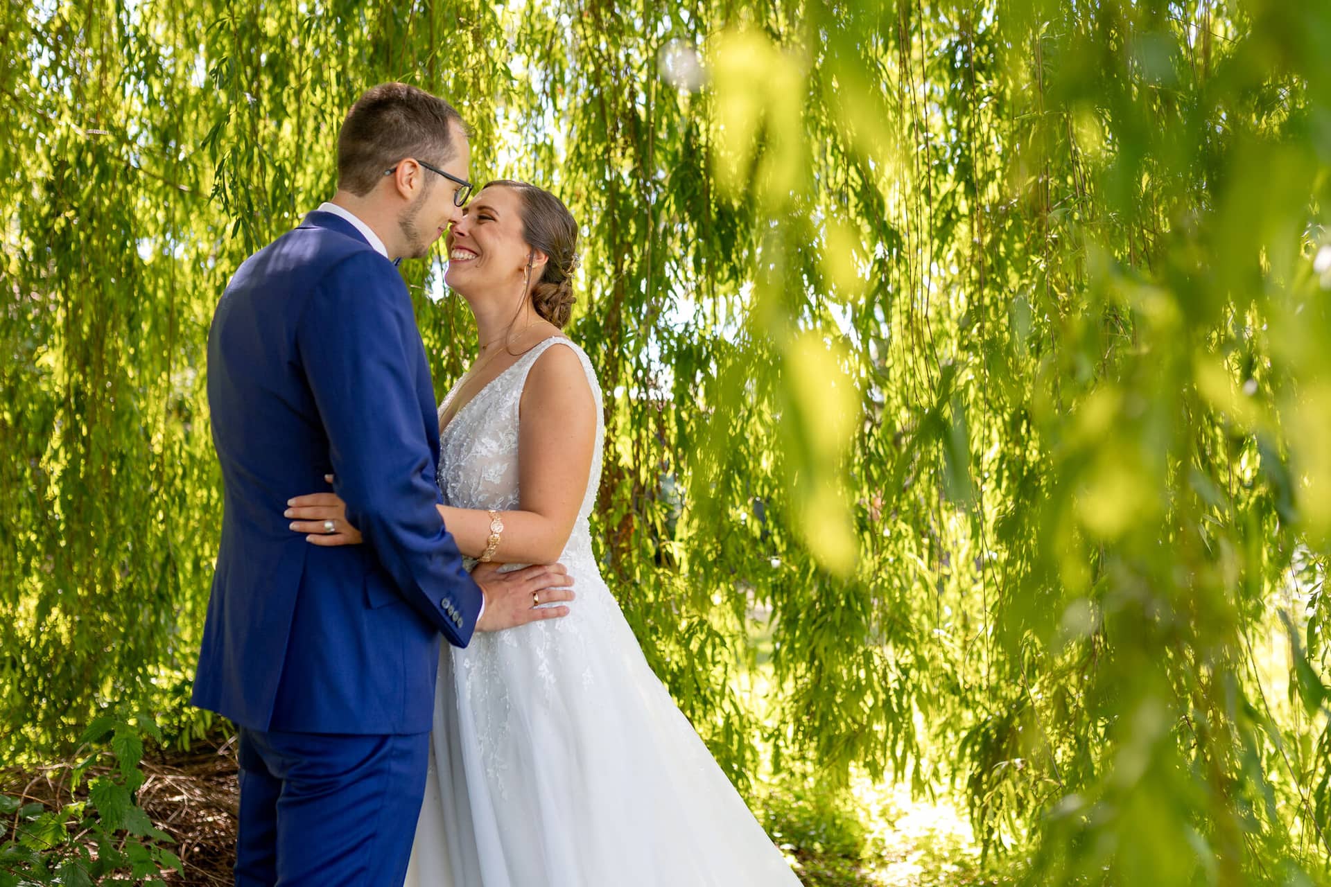 4K8A0416 Hochzeitsfotograf Mutterstadt Grenzhof Heidelberg .ChlXVjYD Brautpaar umarmt sich zärtlich unter einem dichten Weidenbaum im Halbschatten des Grenzhof-Parks