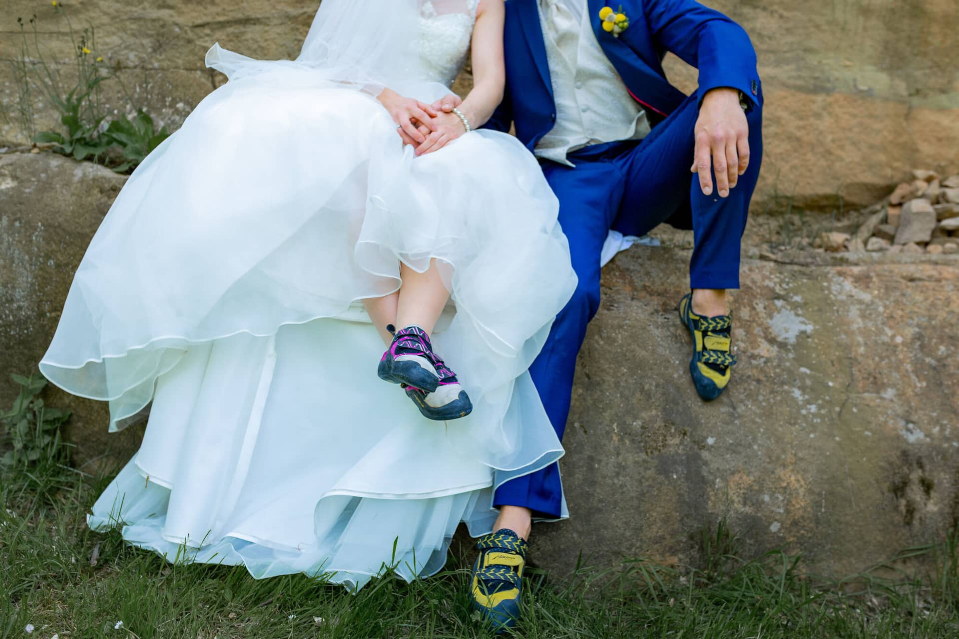 Anne Christian Hochzeitsfotograf Birkenheide Grosskarlbach Bissersheim 00006.DqOaHdyz Braut und Bräutigam sitzen an einem Felsvorsprung, sie im weißen Kleid mit Wanderschuhen, er im blauen Anzug mit gelbem Blumenstrauß
