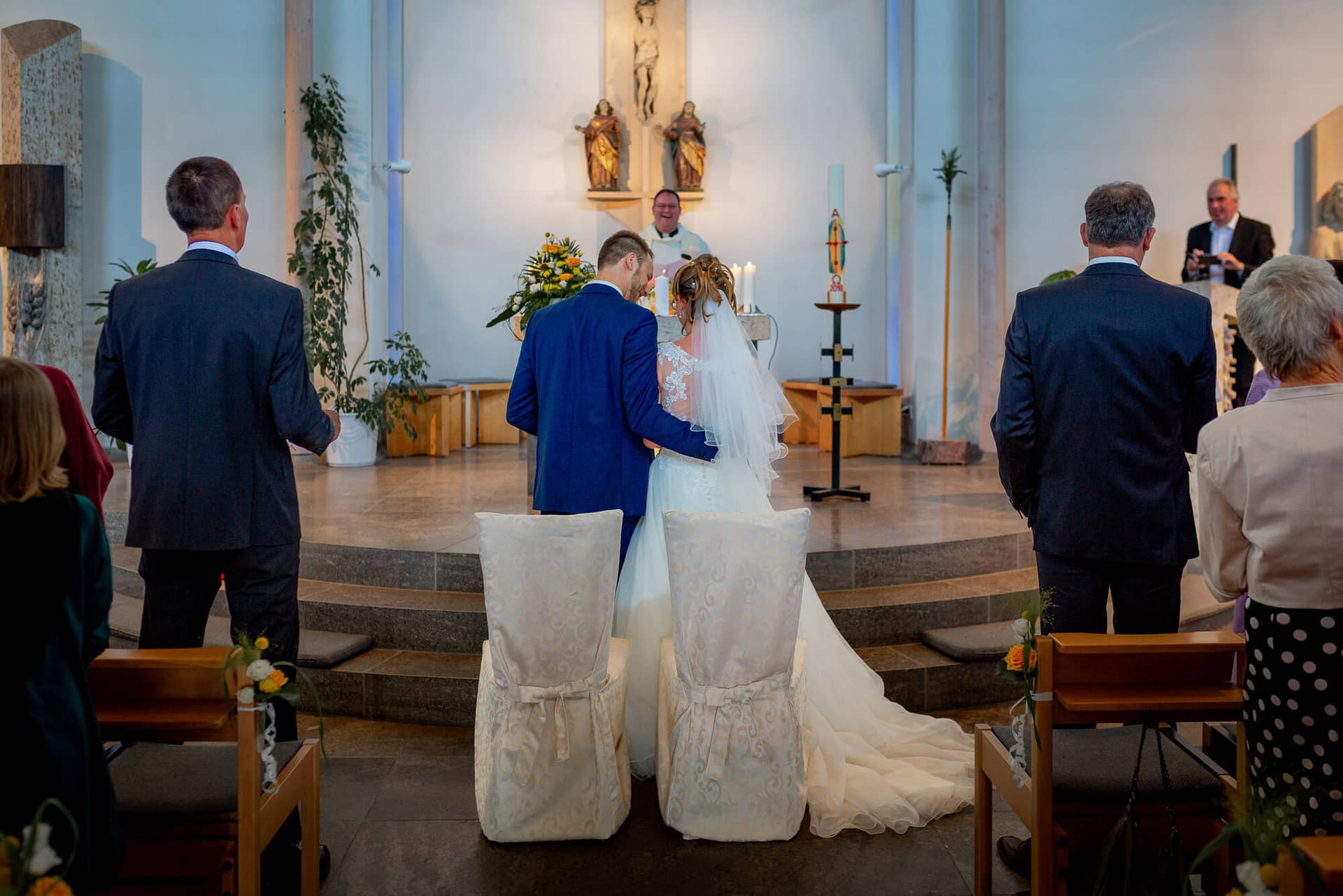 Anne Christian Hochzeitsfotograf Birkenheide Grosskarlbach Bissersheim 00009.DCDBA51X Brautpaar küsst sich beim ersten Kuss in der Kirche, Gäste im Kirchenschiff applaudieren gerührt