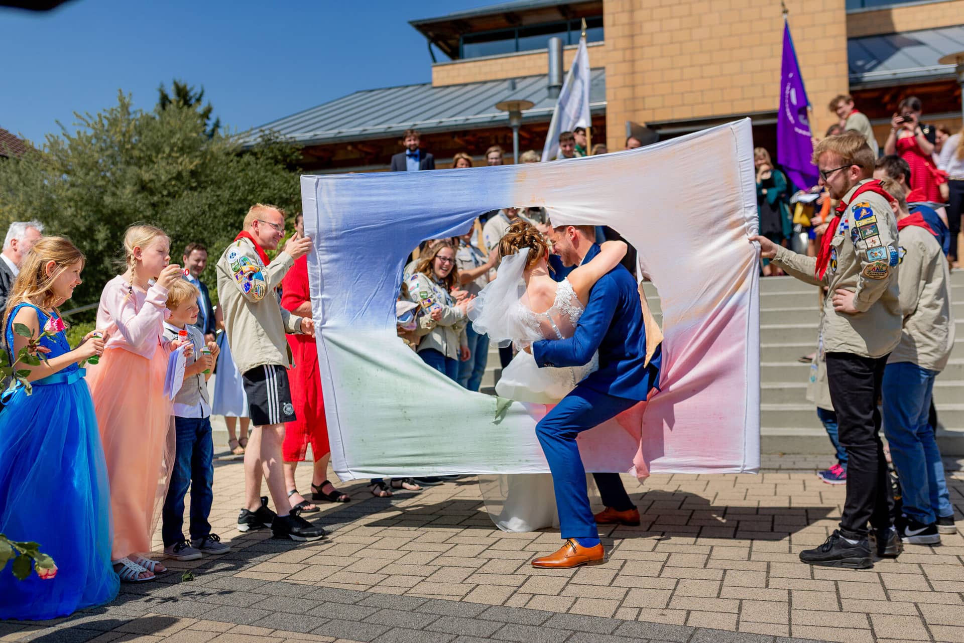 Anne Christian Hochzeitsfotograf Birkenheide Grosskarlbach Bissersheim 00017.C9PnUyO Brautpaar küsst sich leidenschaftlich unter buntem Stoffbanner vor blauem Himmel, Gäste jubeln rundherum