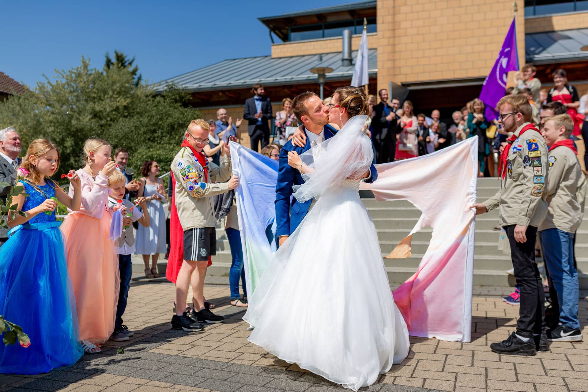 Anne Christian Hochzeitsfotograf Birkenheide Grosskarlbach Bissersheim 00018.CYtgh5vk Braut küsst Bräutigam freudig vor dem Gebäude, während Gäste mit bunten Fahnen jubeln