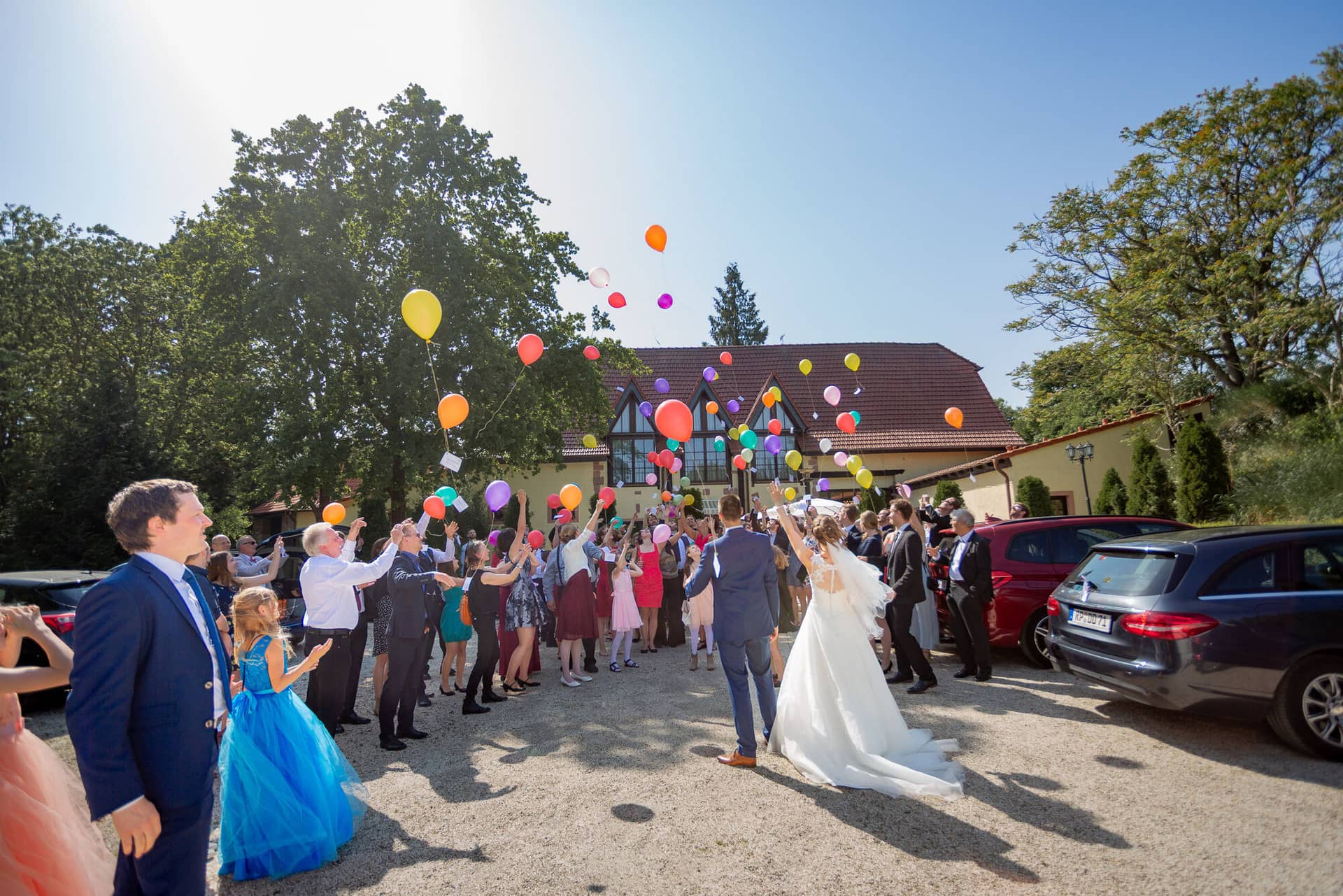 Anne Christian Hochzeitsfotograf Birkenheide Grosskarlbach Bissersheim 00022.CISoNbWP Brautpaar stößt lachend mit bunten Luftballons an, Gäste jubeln rundum beim Sektempfang im Garten