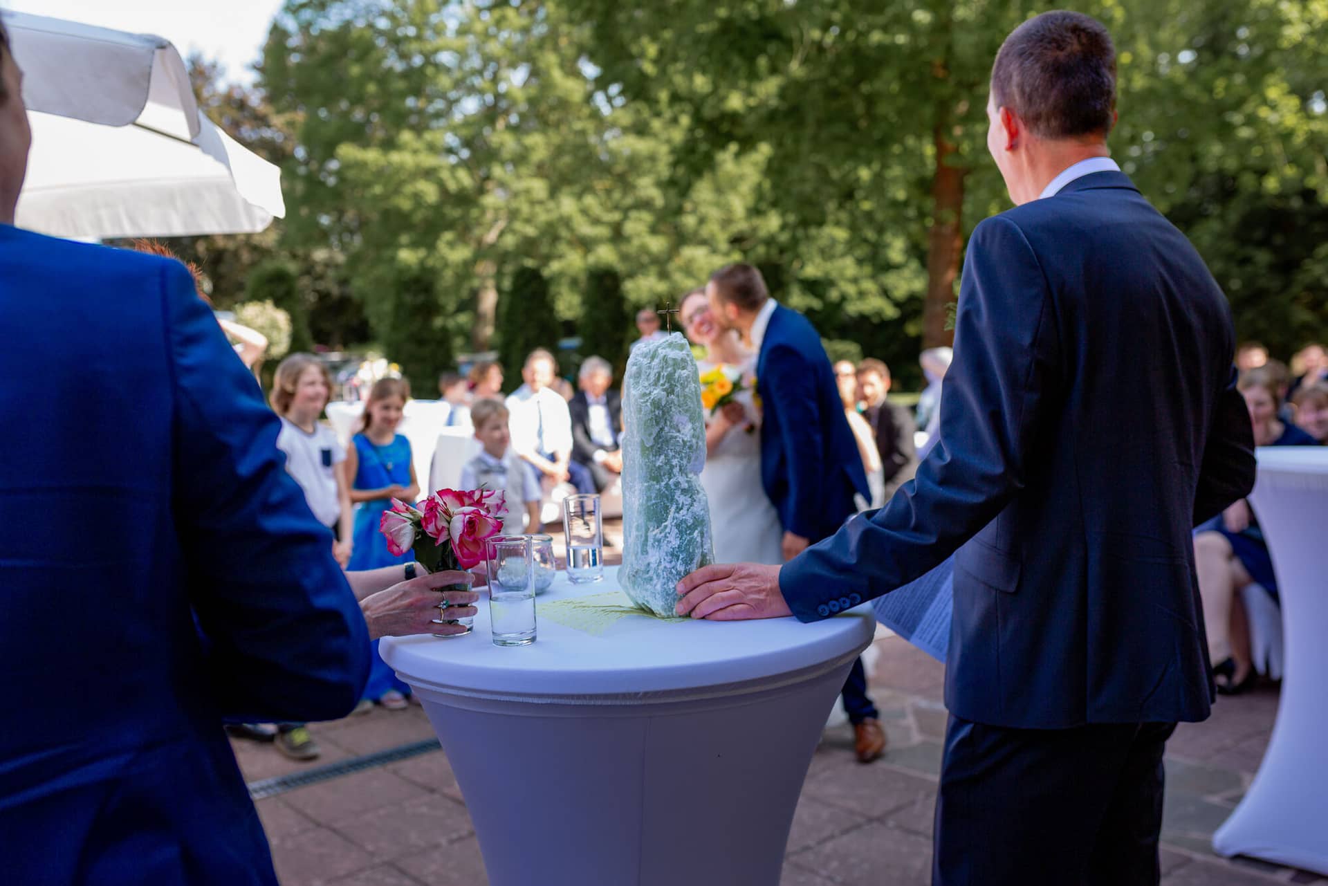 Anne Christian Hochzeitsfotograf Birkenheide Grosskarlbach Bissersheim 00023.C E3gRkK Trauzeugenleiter führt das Brautpaar beim Hochzeitsritual vor einem weißen Blumentisch mit Grünpflanzentuch