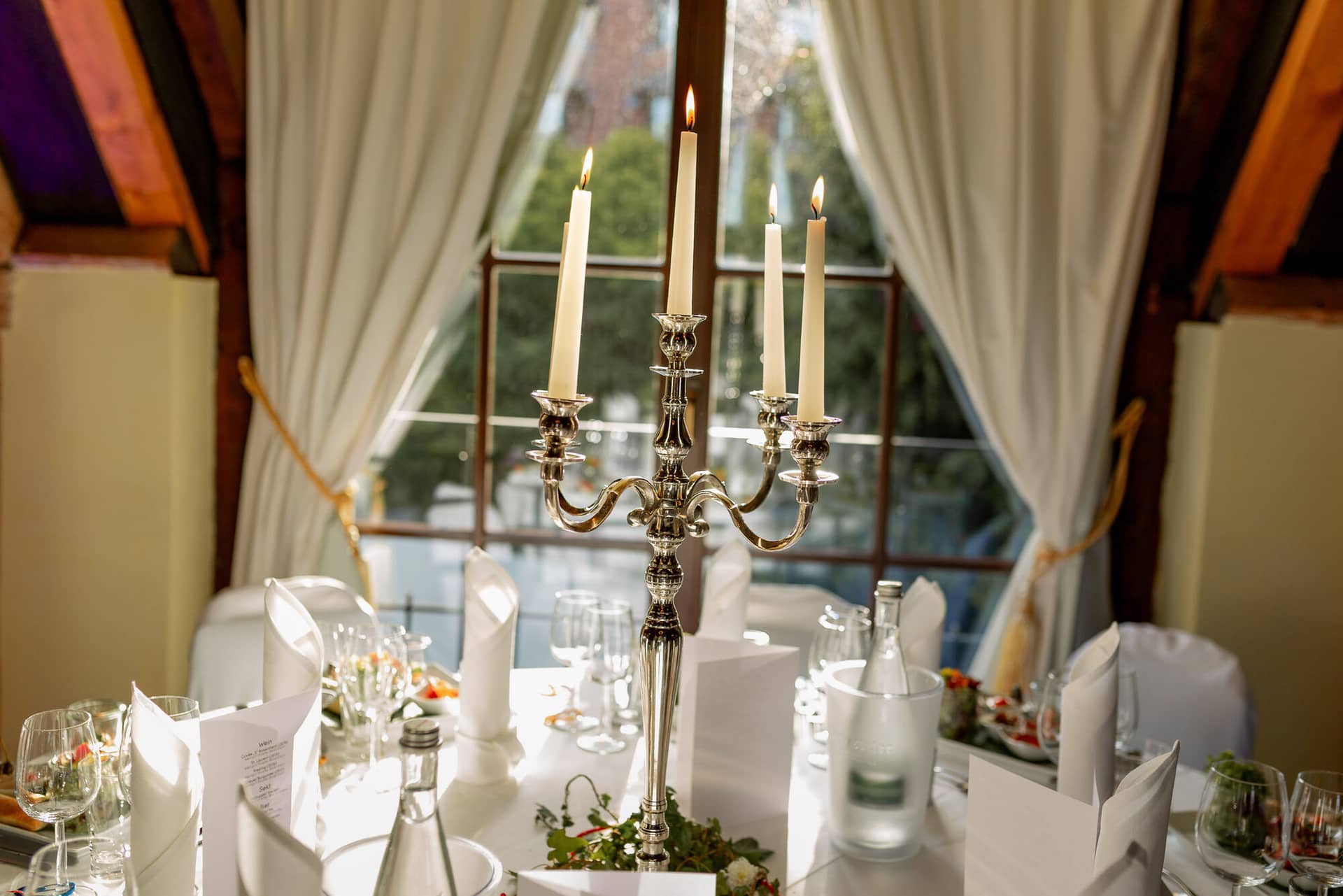Anne Christian Hochzeitsfotograf Birkenheide Grosskarlbach Bissersheim 00033.D3iWTkt3 Silberne Kerzenleuchter mit Kerzen auf gedecktem Hochzeitstisch mit Menükarte vor dem Saalfenster