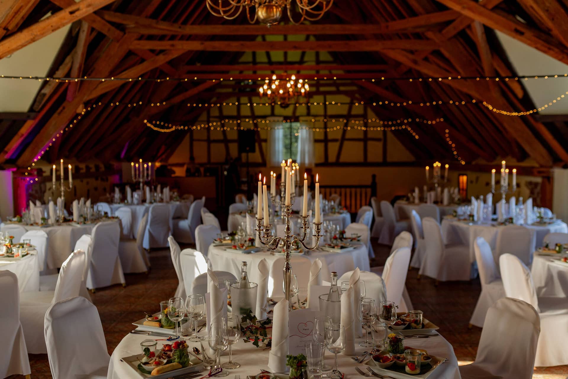 Anne Christian Hochzeitsfotograf Birkenheide Grosskarlbach Bissersheim 00034.Dml3AIhJ Hochzeitsdinner in strohgedecktem Saal mit Kerzen, weißen Tischdecken und Gästen an langen Tafeln