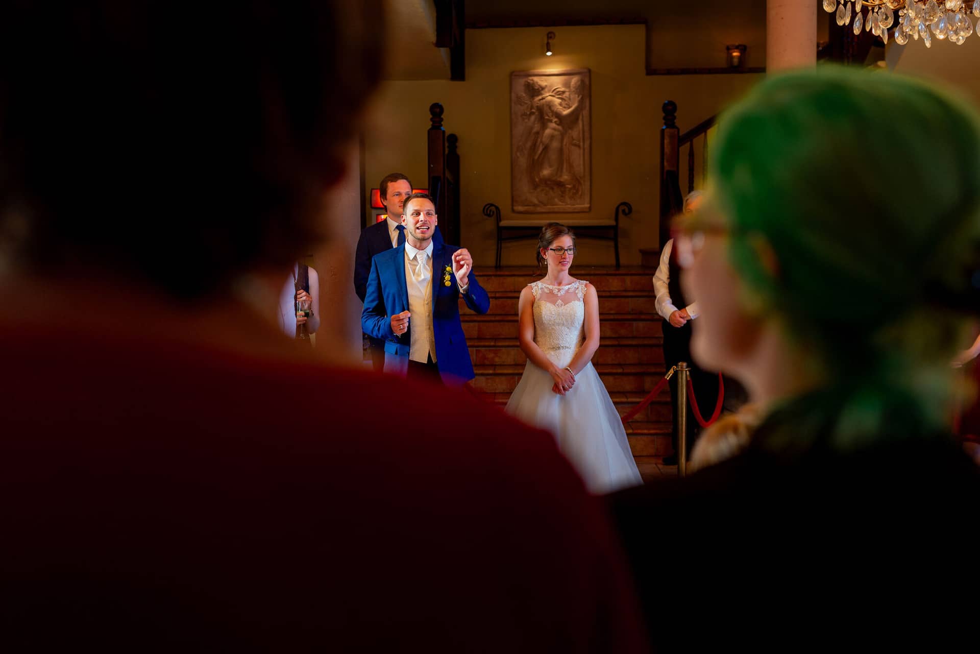 Anne Christian Hochzeitsfotograf Birkenheide Grosskarlbach Bissersheim 00035.CsxM71IO Bräutigam steht elegant in historischem Saal mit Treppe und Kristalllüstern, Braut tritt im Hintergrund ein