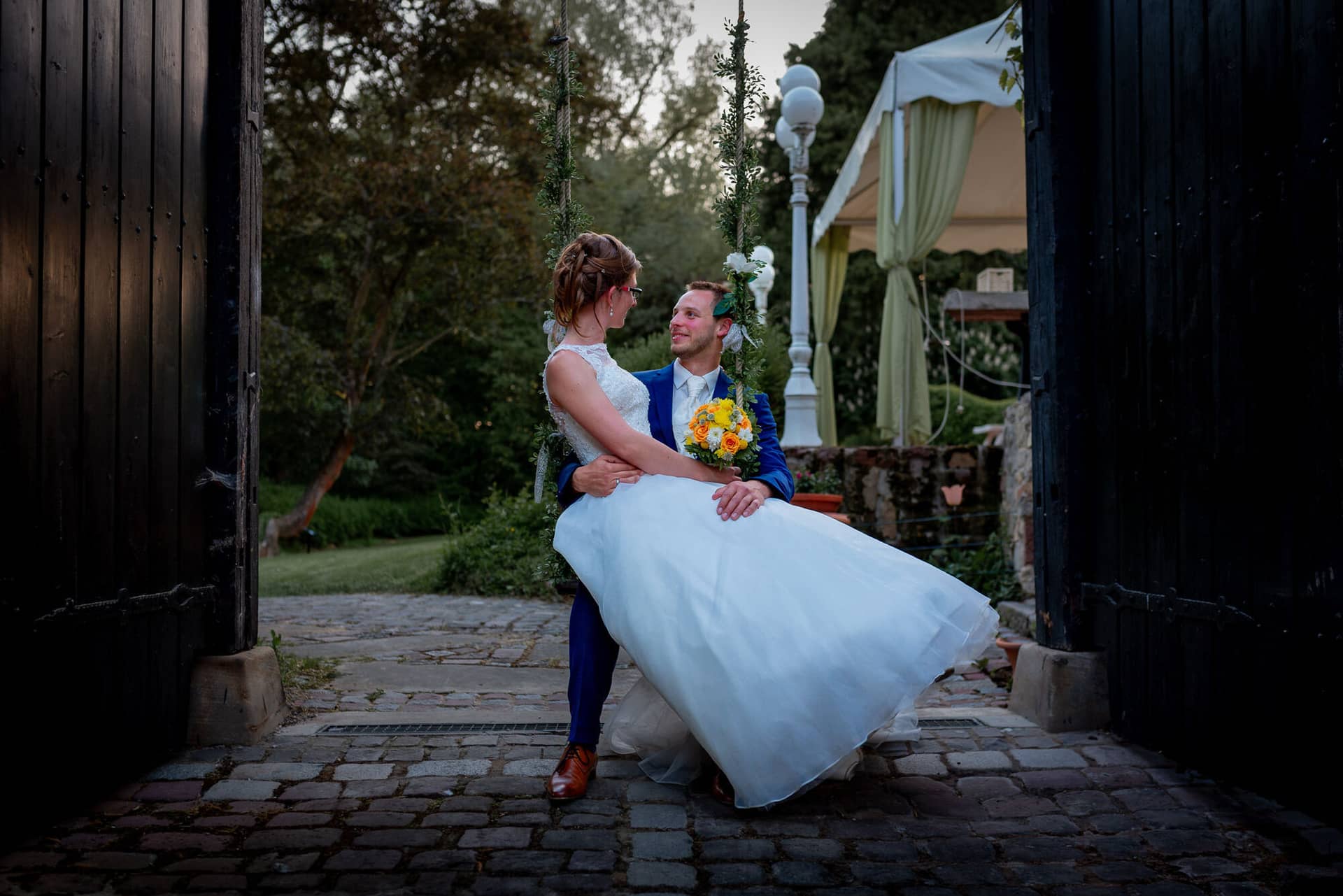 Anne Christian Hochzeitsfotograf Birkenheide Grosskarlbach Bissersheim 00040.B5NTC83p Brautpaar sitzt eng umschlungen an einem Holzzaun unter grünem Pavillon mit gelben Blüten