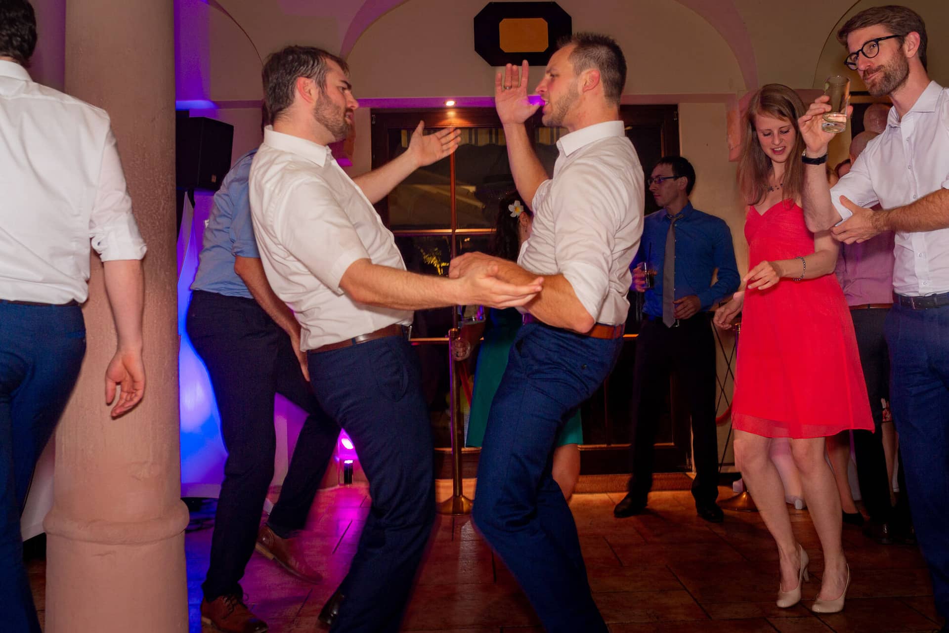 Anne Christian Hochzeitsfotograf Birkenheide Grosskarlbach Bissersheim 00048.XoylODn8 Zwei Männer tanzen begeistert auf der Hochzeitsfeier, eine Frau beobachtet sie lächelnd