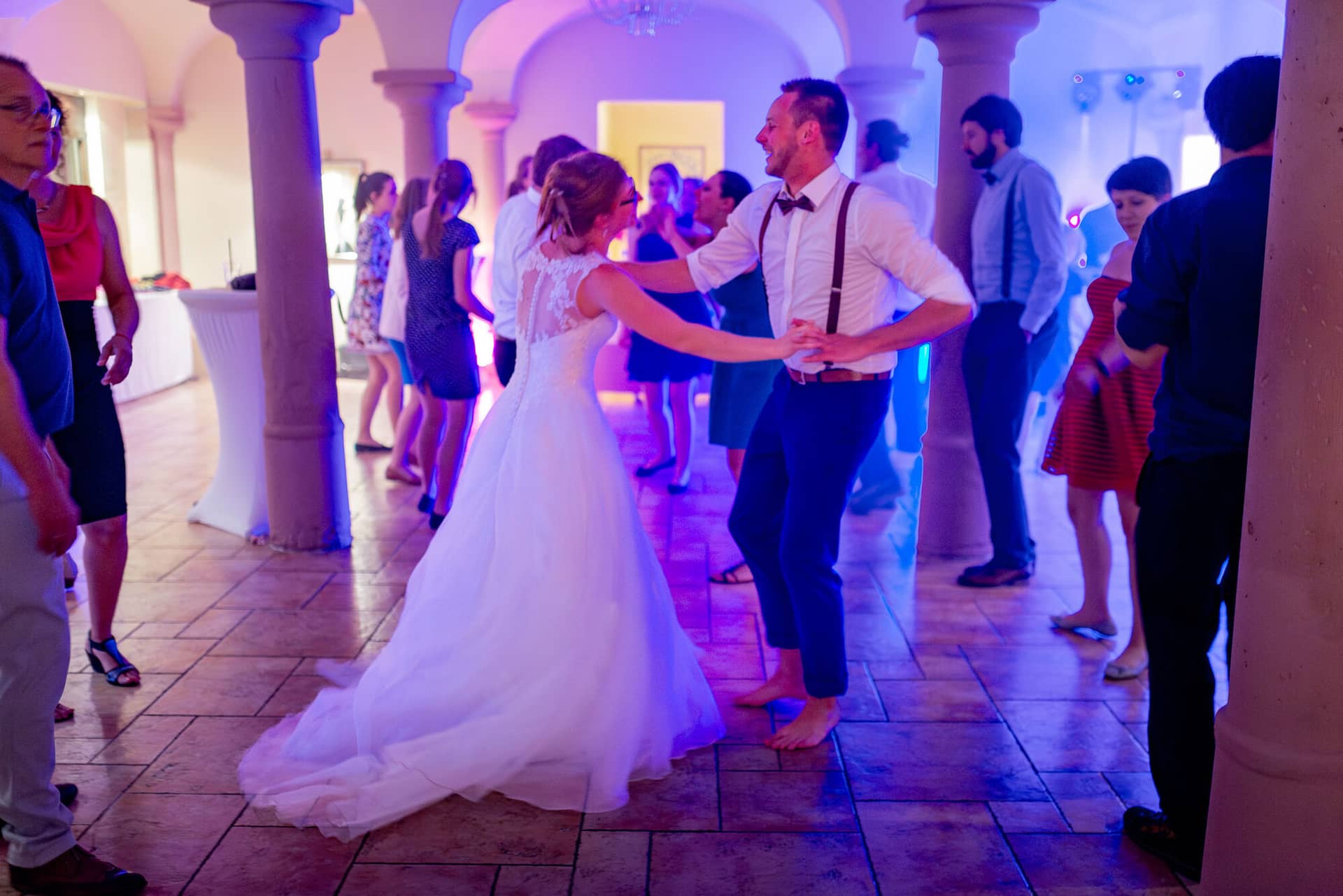 Anne Christian Hochzeitsfotograf Birkenheide Grosskarlbach Bissersheim 00053.6HLDysM8 Brautpaar tanzt ausgelassen im blauen Scheinwerferlicht in Saal mit Säulen bei der Hochzeitsparty