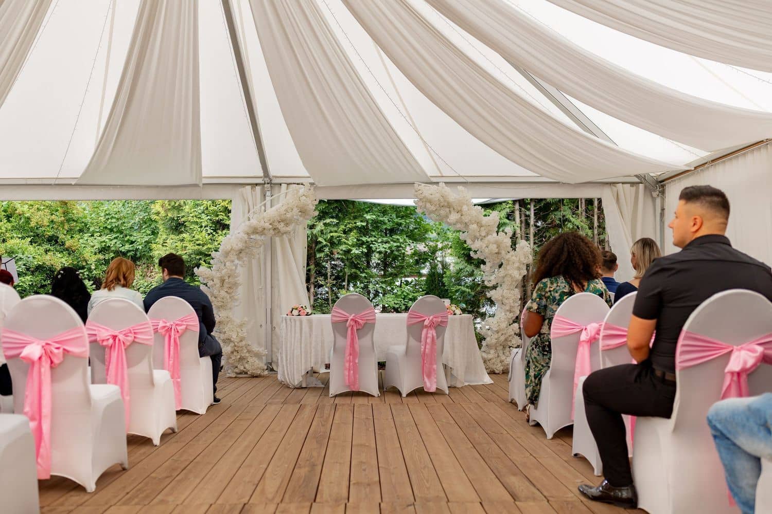 Brautpaarshooting Weinheim Schlossgarten Heidelberg Freie Trauung 4K8A5984 7b30ca09 1500.DIocdl Q Hochzeitsgäste sitzen unter weißem Zelt bei der freien Trauung, weißer Blumenbogen als Altar, Bäume im Hintergrund