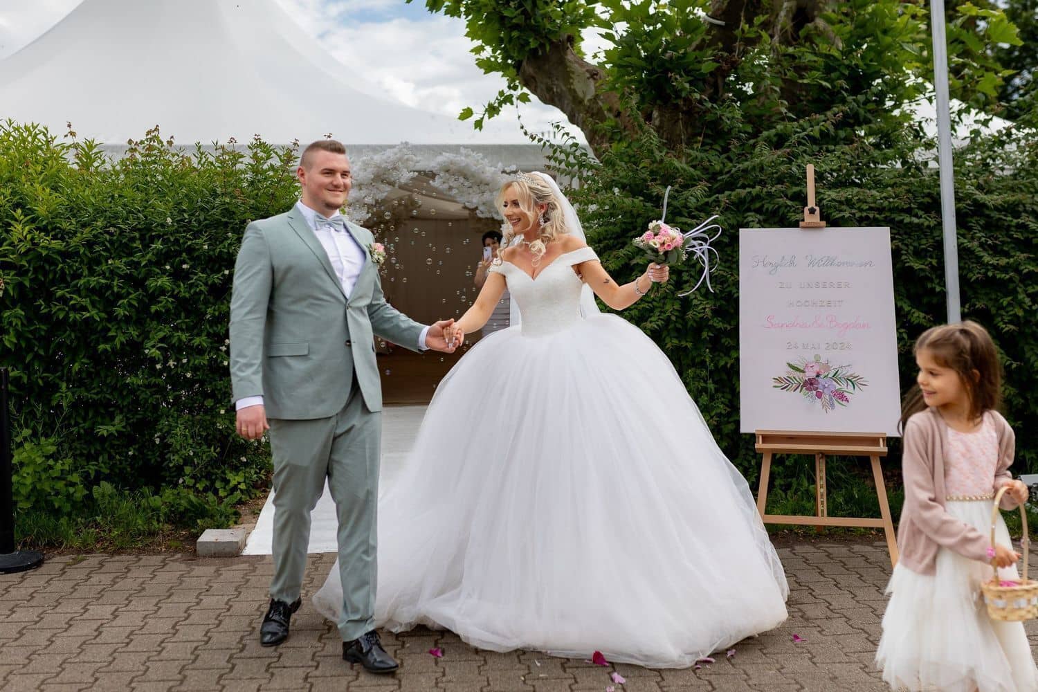 Brautpaarshooting Weinheim Schlossgarten Heidelberg Freie Trauung 4K8A6952 168042a6 1500.B8LSdj8p Brautpaar geht strahlend durch eine Seifenblasen-Dusche, kleines Mädchen mit Blume begleitet das Paar