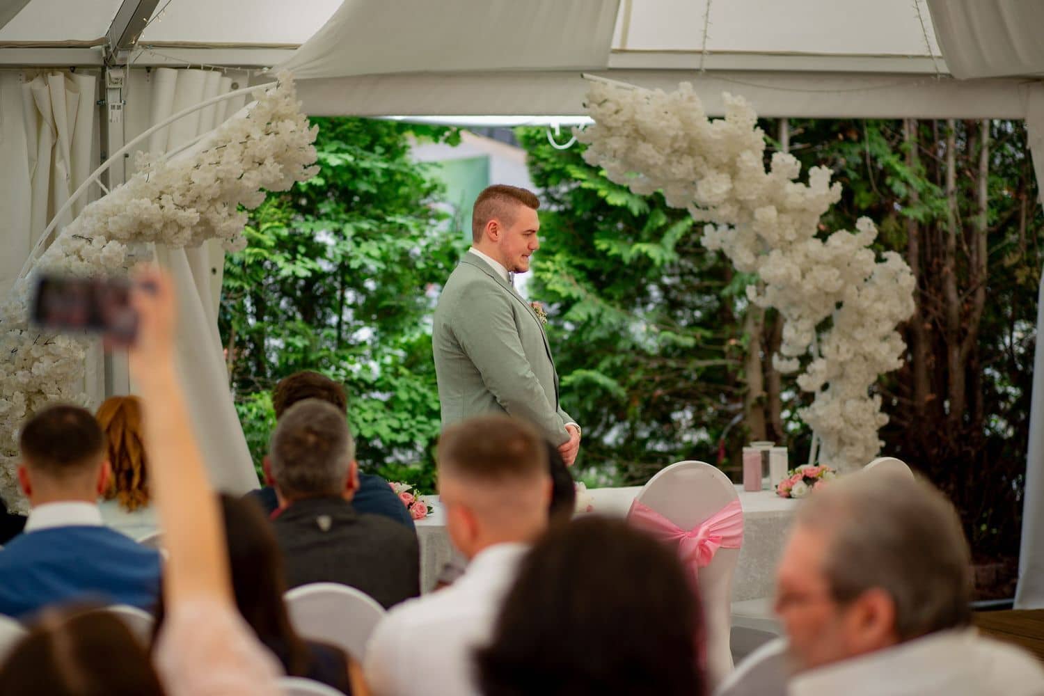 Brautpaarshooting Weinheim Schlossgarten Heidelberg Freie Trauung MK3 4649 6021db14 1500.D4LyEdIB Bräutigam wartet am weißen Blumenbogen beim Traualtar im Zelt, Hochzeitsgäste sitzen auf Stühlen dahinter