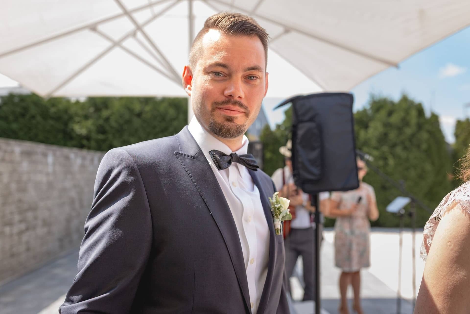 Hochzeit Schwetzingen Hochzeitsfotograf Heiraten Miio MK3 7221.DOODBFoH Gutgelaunter Gast im dunkelblauen Anzug mit Fliege, Braut und Bräutigam im Hintergrund beim Empfang