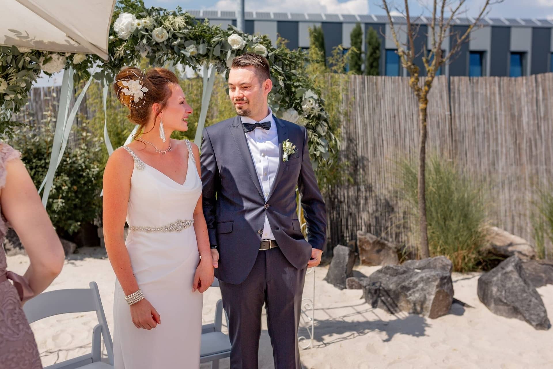 Hochzeit Schwetzingen Hochzeitsfotograf Heiraten Miio MK3 7284.C8kp9MY6 Brautpaar steht vereint unter dem weißen Blumenbogen, Sanduntergrund des Miio Schwetzingen