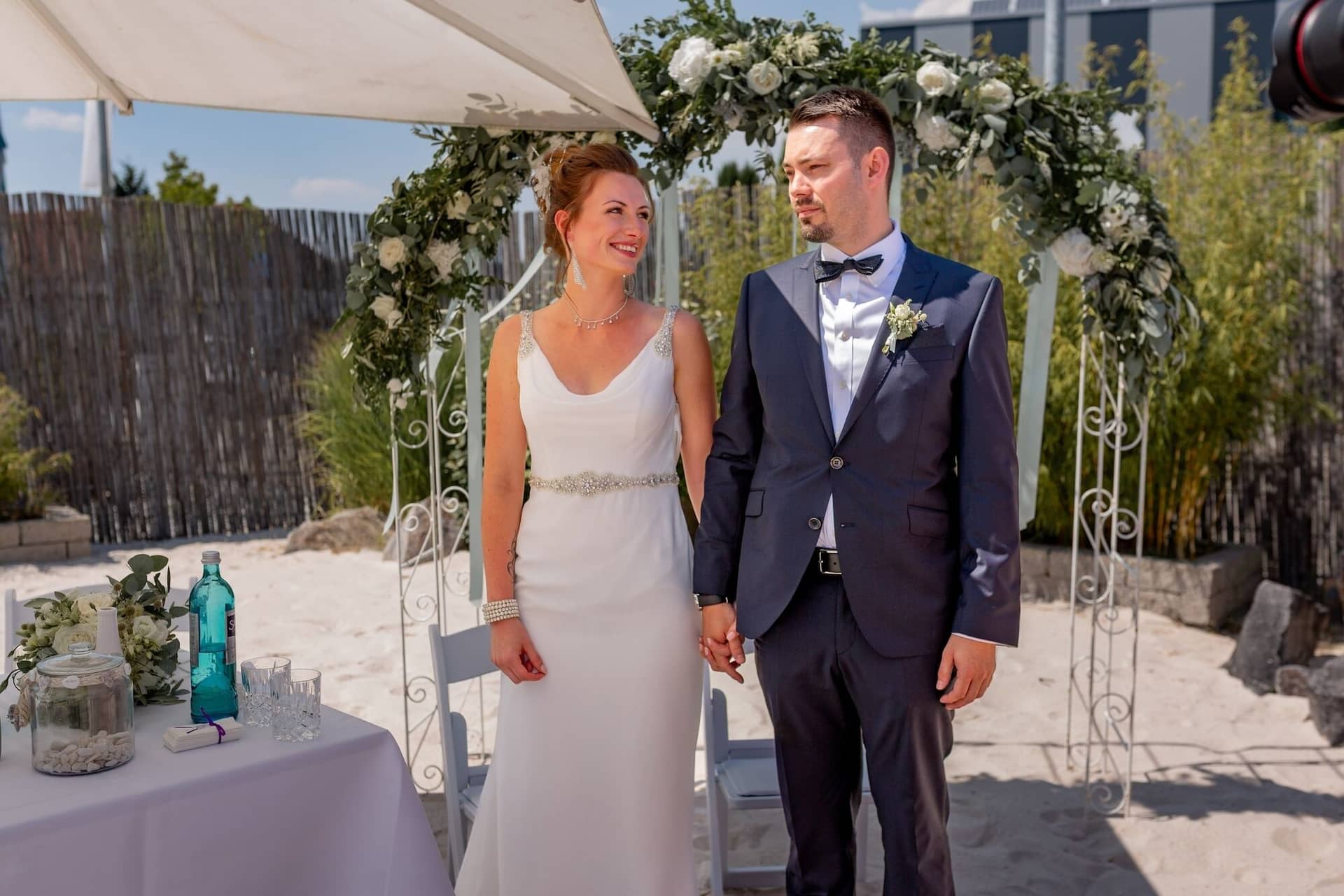 Hochzeit Schwetzingen Hochzeitsfotograf Heiraten Miio MK3 7389.BFE9gx1x Glückliches Brautpaar küsst sich nach der Trauung vor dem weißen Blumenbogen, Sonnenstimmung