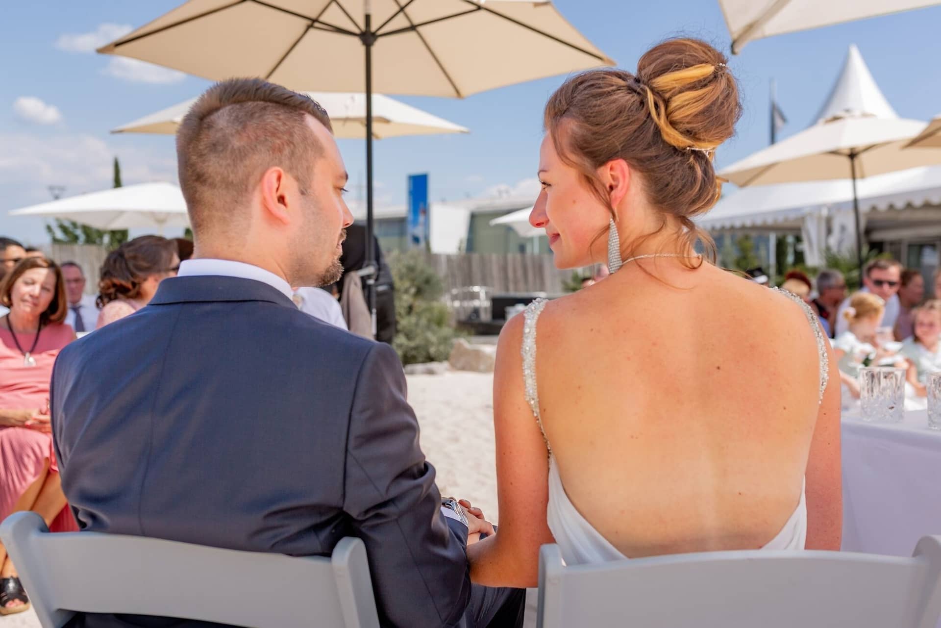 Hochzeit Schwetzingen Hochzeitsfotograf Heiraten Miio MK3 7554.CIcI UAU Brautpaar sitzt beim Empfang auf dem Sandstrand, umgeben von Gästen mit Sonnenschirmen