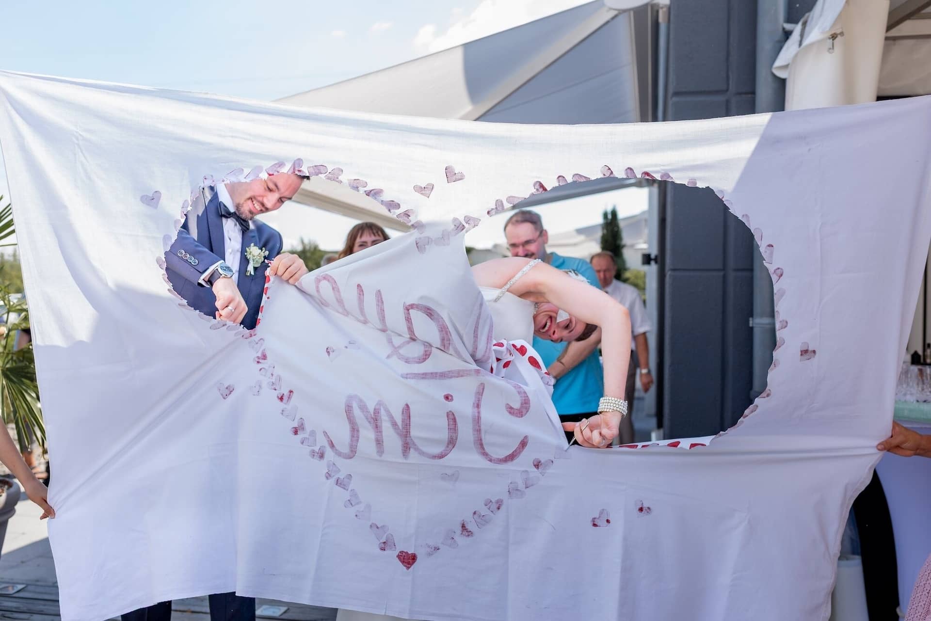 Hochzeit Schwetzingen Hochzeitsfotograf Heiraten Miio MK3 8029.BS3Uh7hc Bräutigam hält mit Braut ein großes "SMILE"-Tuch mit Herzchen, Gäste schauen lachend zu