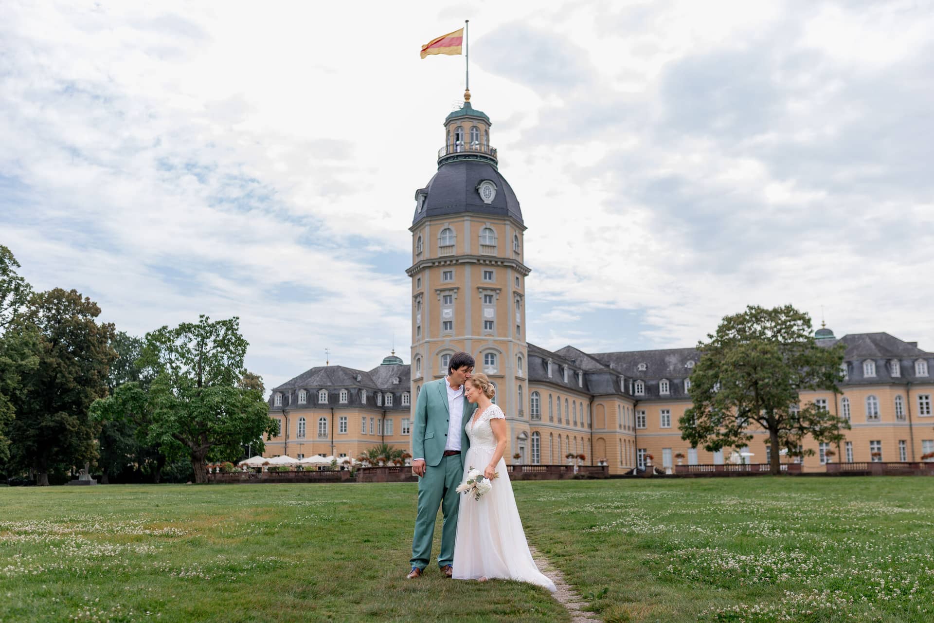 Hochzeitsfotograf Karlsruhe Daniela Matthias 20230819 13 54 49 4K8A5221.BRxDM0ZM Brautpaar in weißem Kleid und grünem Anzug vor dem imposanten Karlsruher Schloss mit hoher Turmspitze