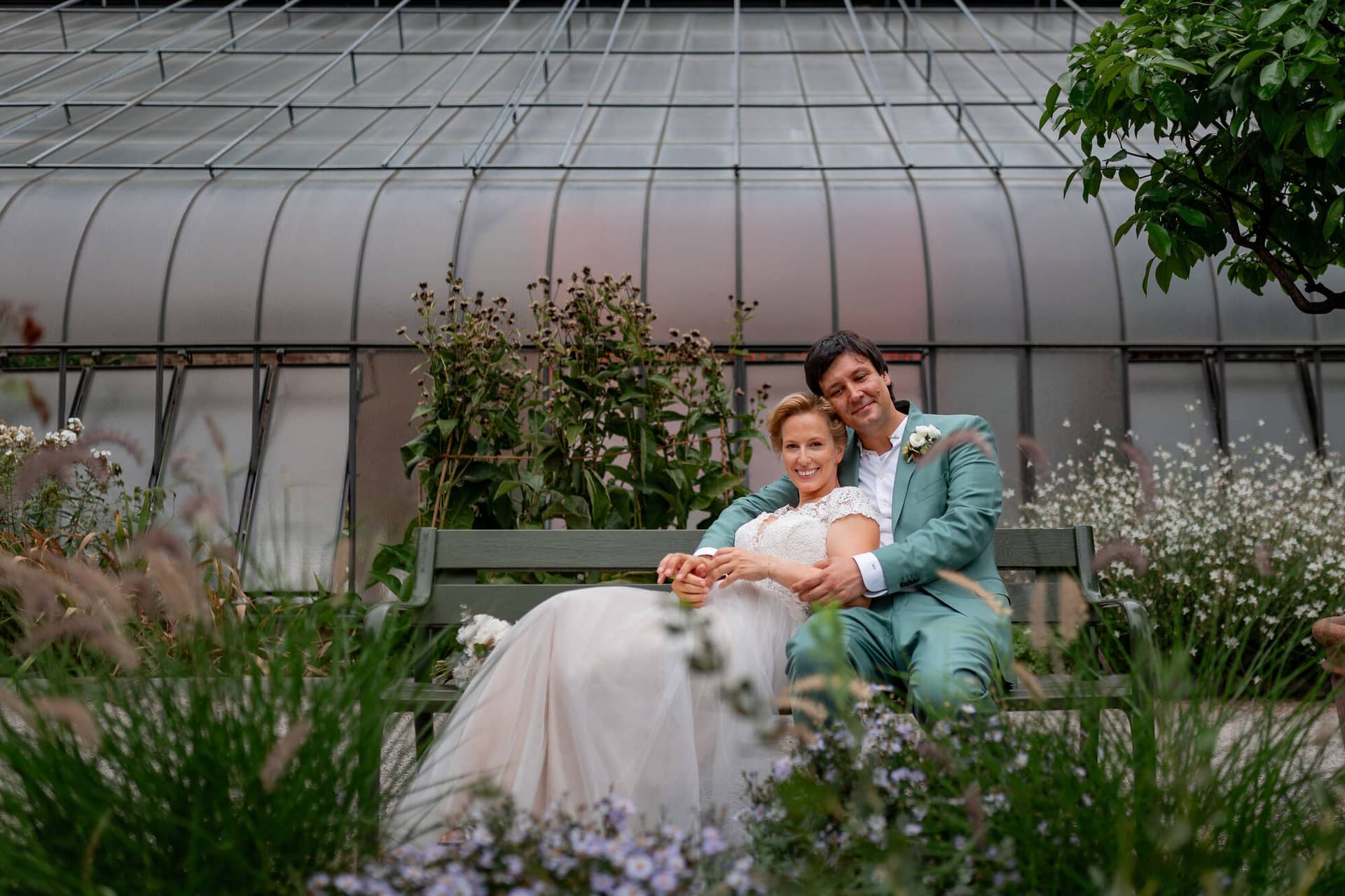 Hochzeitsfotograf Karlsruhe Daniela Matthias 20230819 14 21 39 4K8A5698.B9BZXlGo Brautpaar sitzt eng aneinandergeschmiegt auf einer Bank im Gewächshaus, umgeben von üppig blühenden Pflanzen