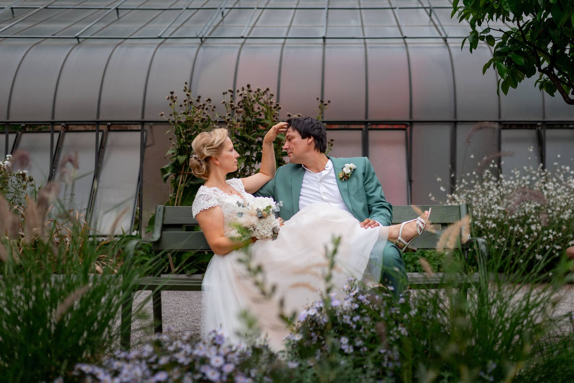 Hochzeitsfotograf Karlsruhe Daniela Matthias 20230819 14 22 30 4K8A5785.BRuOta5S Brautpaar sitzt auf Bank in blühendem Garten mit Lavendel und Wildblumen, Glasdach des Gewächshauses darüber