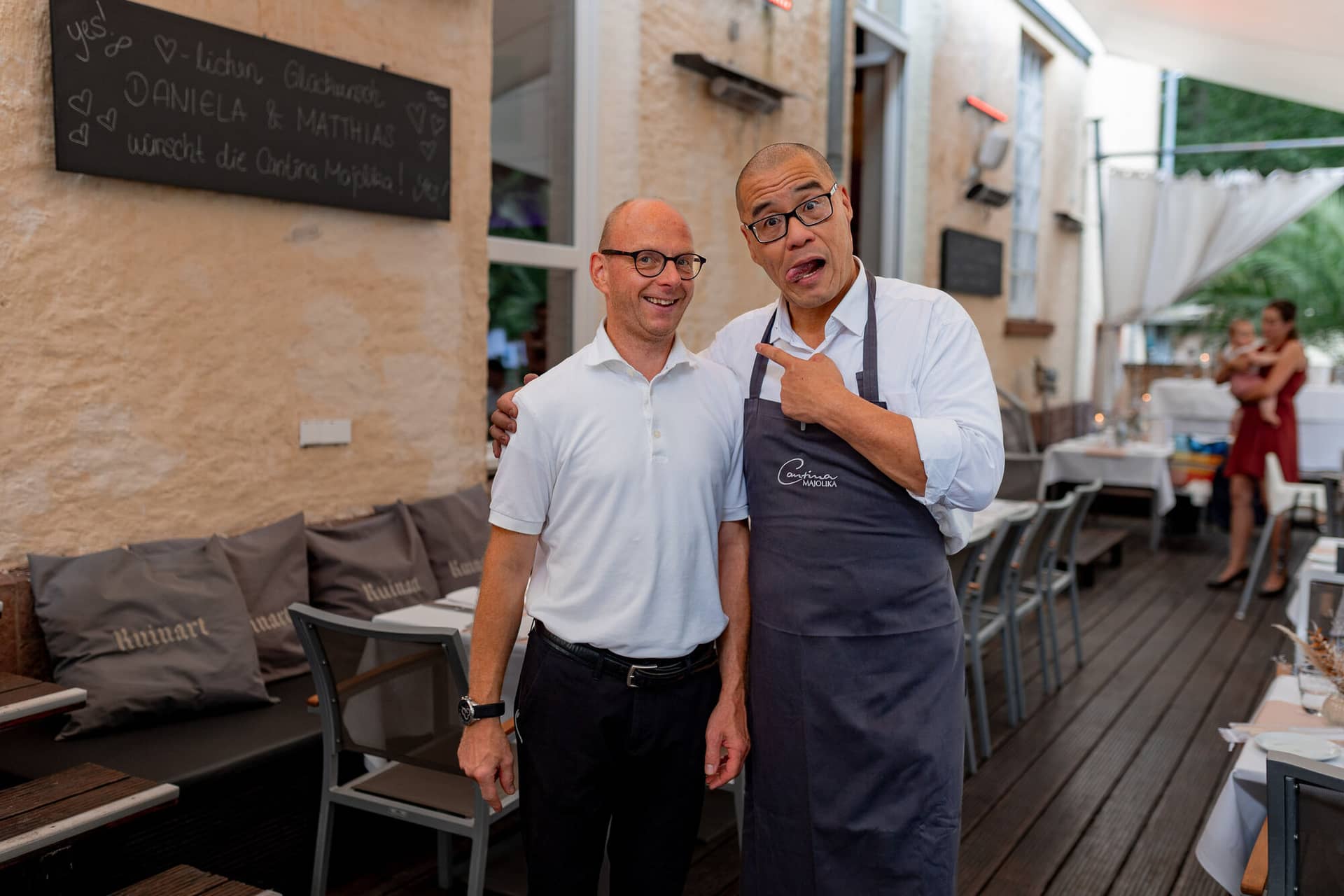 Hochzeitsfotograf Karlsruhe Daniela Matthias 20230819 18 06 14 4K8A9586.BmxYpzcf Brautpaar lacht herzlich mit Herz-Schild „Daniela & Matthias" auf der Terrasse der Cantina Majolika