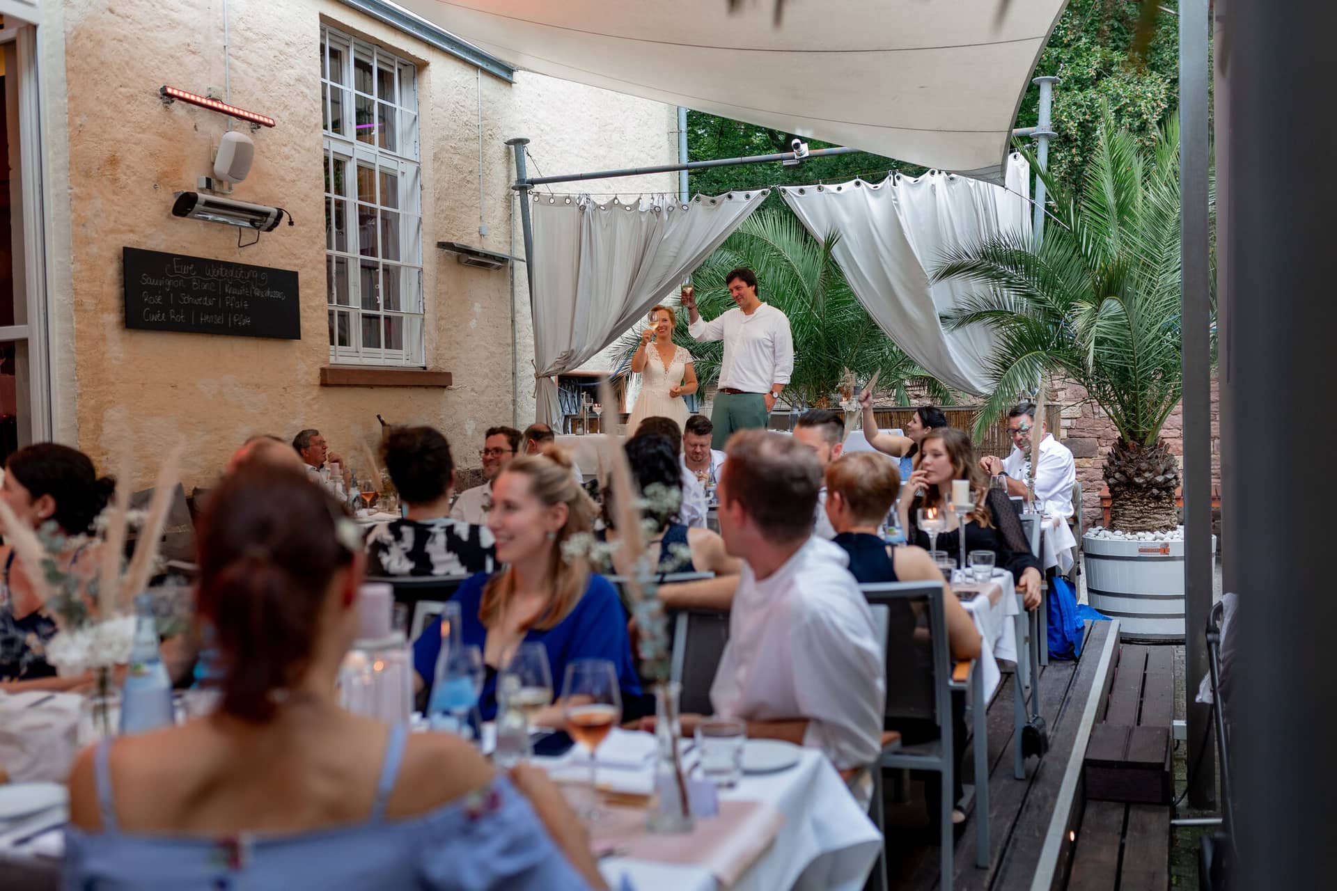 Hochzeitsfotograf Karlsruhe Daniela Matthias 20230819 18 37 09 4K8A9745. TLyE2OO Hochzeitsgesellschaft beim Abendessen auf der Terrasse, Brautpaar erhebt die Gläser zu einem Toast
