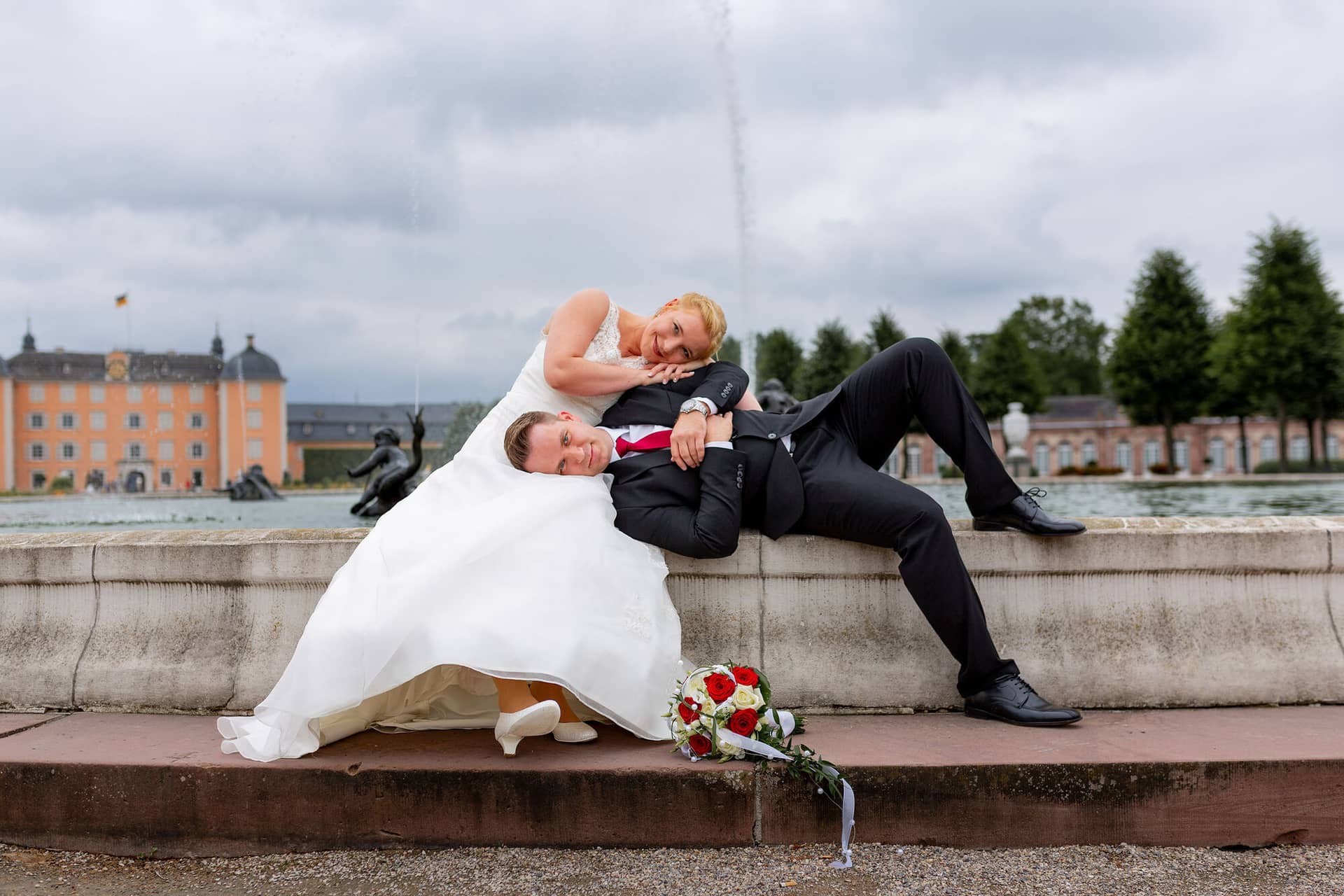 Hochzeitsfotograf Schwetzingen Schlossgarten Brautpaarshooting 20170812 14 03 16 IMG 0144.Ype0Z4Hf Brautpaar liegt entspannt auf einer roten Steinmauer, Schloss Schwetzingen und Brunnen im Hintergrund