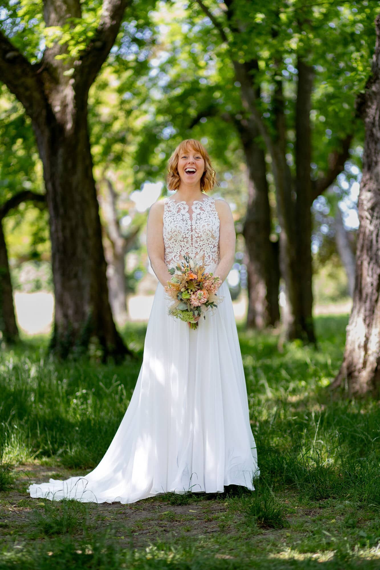 Hochzeitsfotograf Worms Frankenthal Hoftgut Petersau Brautpaarshooting 20230603 13 39 09 4K8A3309.4Fz7L3Hw Braut in weißem Spitzenkleid mit Boho-Blumenstrauß lacht strahlend im Waldbereich des Schlossgartens