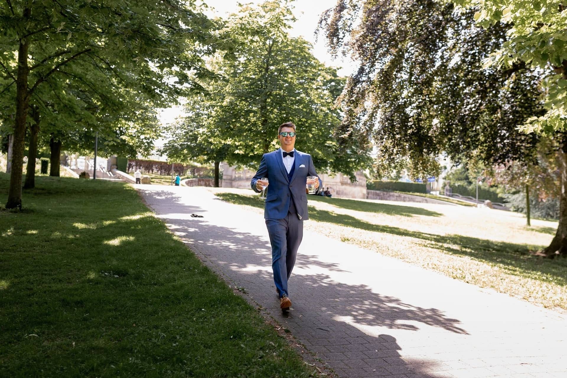 Standesamtliche Trauung Mutterstadt Altes Rathaus Bad Duerkheim Schlossgarten IMG 3361.1k8udFW7 Bräutigam im dunkelblauen Anzug und Sonnenbrille schlendert mit Glas durch den Kurpark Bad Dürkheim