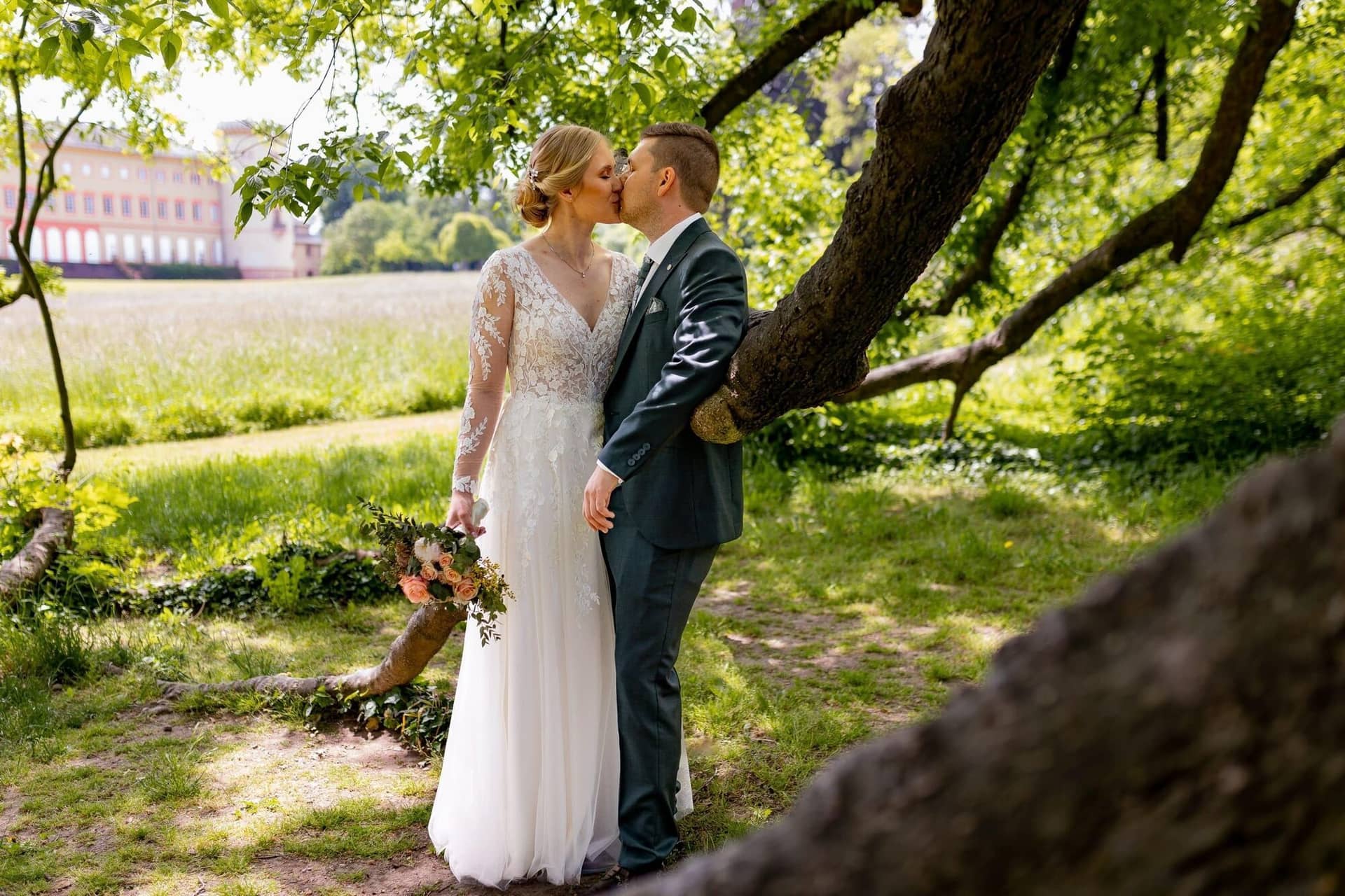 Verena Marius Hochzeitsfotograf Fotograf Worms Mettenheim 01.CV8EqoCJ Brautpaar küsst sich leidenschaftlich unter einem alten Baum mit Schloss Herrnsheim im Hintergrund
