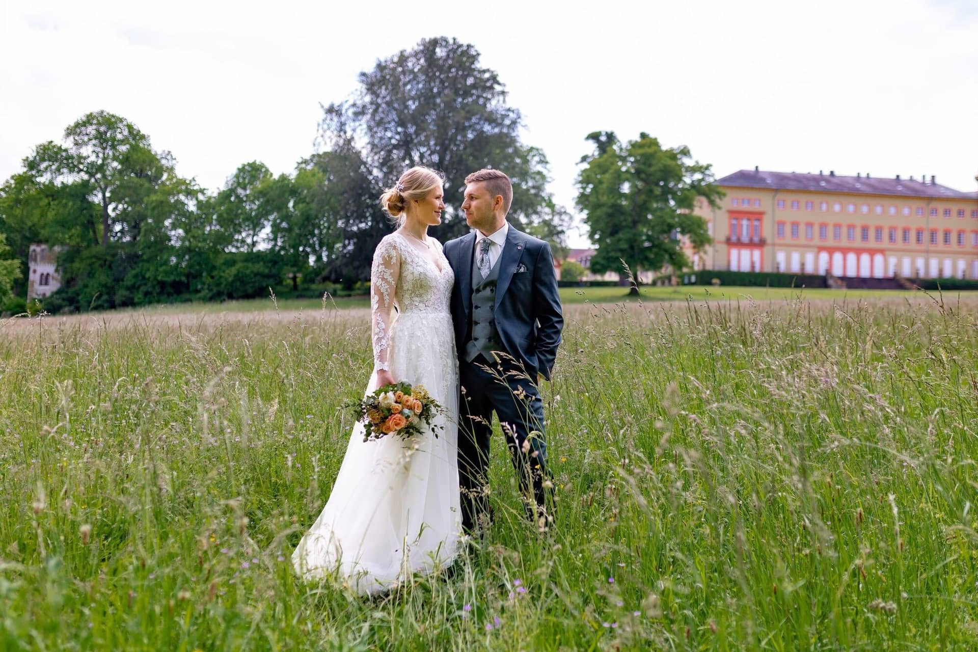 Verena Marius Hochzeitsfotograf Fotograf Worms Mettenheim 02.LOObUHRn Brautpaar steht Hand in Hand im hohen Gras vor dem gelb-orangen Schloss Herrnsheim, Bräutigam im grauen Anzug, Braut mit Schleier