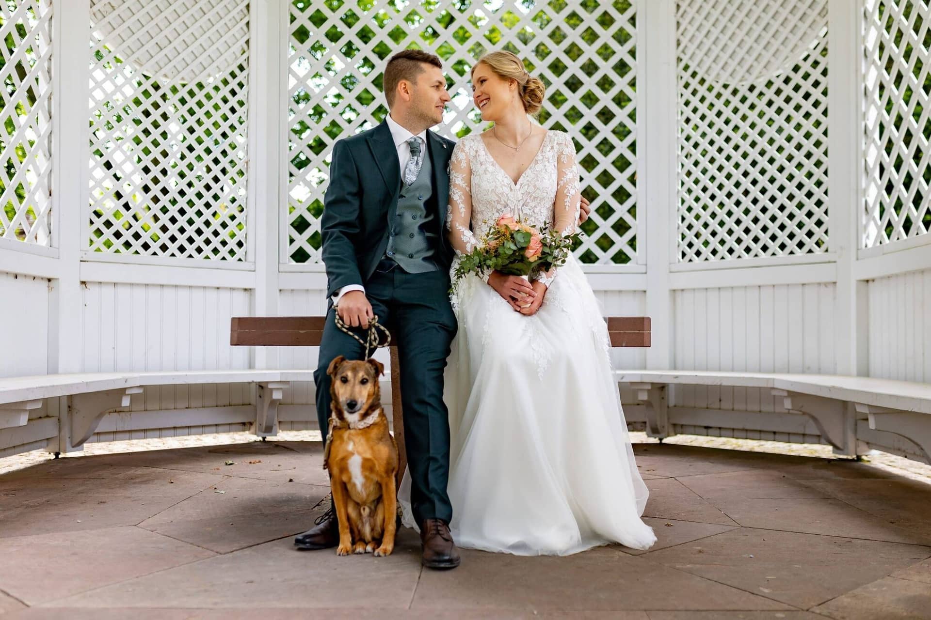Verena Marius Hochzeitsfotograf Fotograf Worms Mettenheim 03.Ul7T53gU Brautpaar sitzt lächelnd mit ihrem braunen Hund in einer weißen Pergola im Schlossgarten Herrnsheim