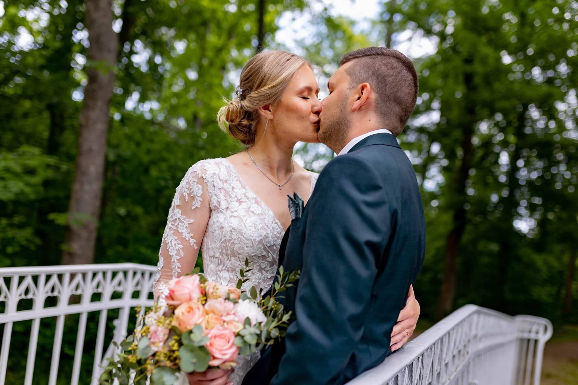 Verena Marius Hochzeitsfotograf Fotograf Worms Mettenheim 04.Cwd4quM9 Brautpaar küsst sich auf einer weißen Bank im Waldbereich des Schlossgartens, Braut hält Boho-Blumenstrauß in der Hand