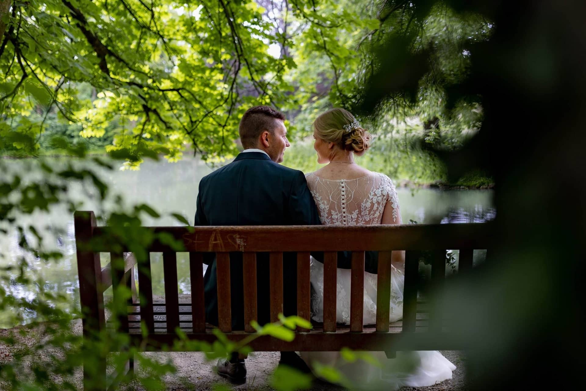 Verena Marius Hochzeitsfotograf Fotograf Worms Mettenheim 05.DGuRkx85 Brautpaar sitzt auf einer Bank am Teich und blickt sich liebevoll an, Bräutigam im dunkelblauen Anzug, ruhige Gartenatmosphäre